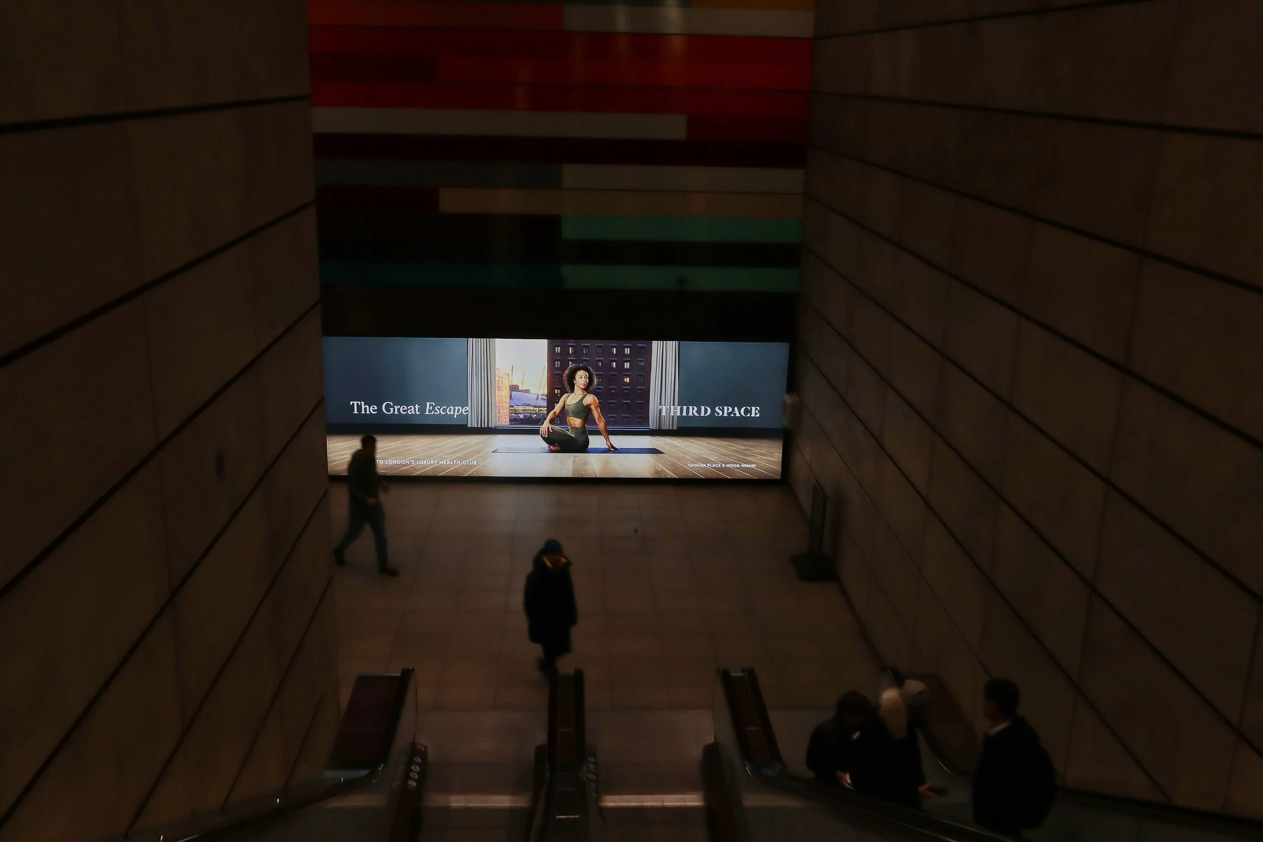 People descending escalators in a dimly lit hallway toward a large digital advertisement featuring a woman practicing yoga with the text "The Great Escape" and "Third Space."