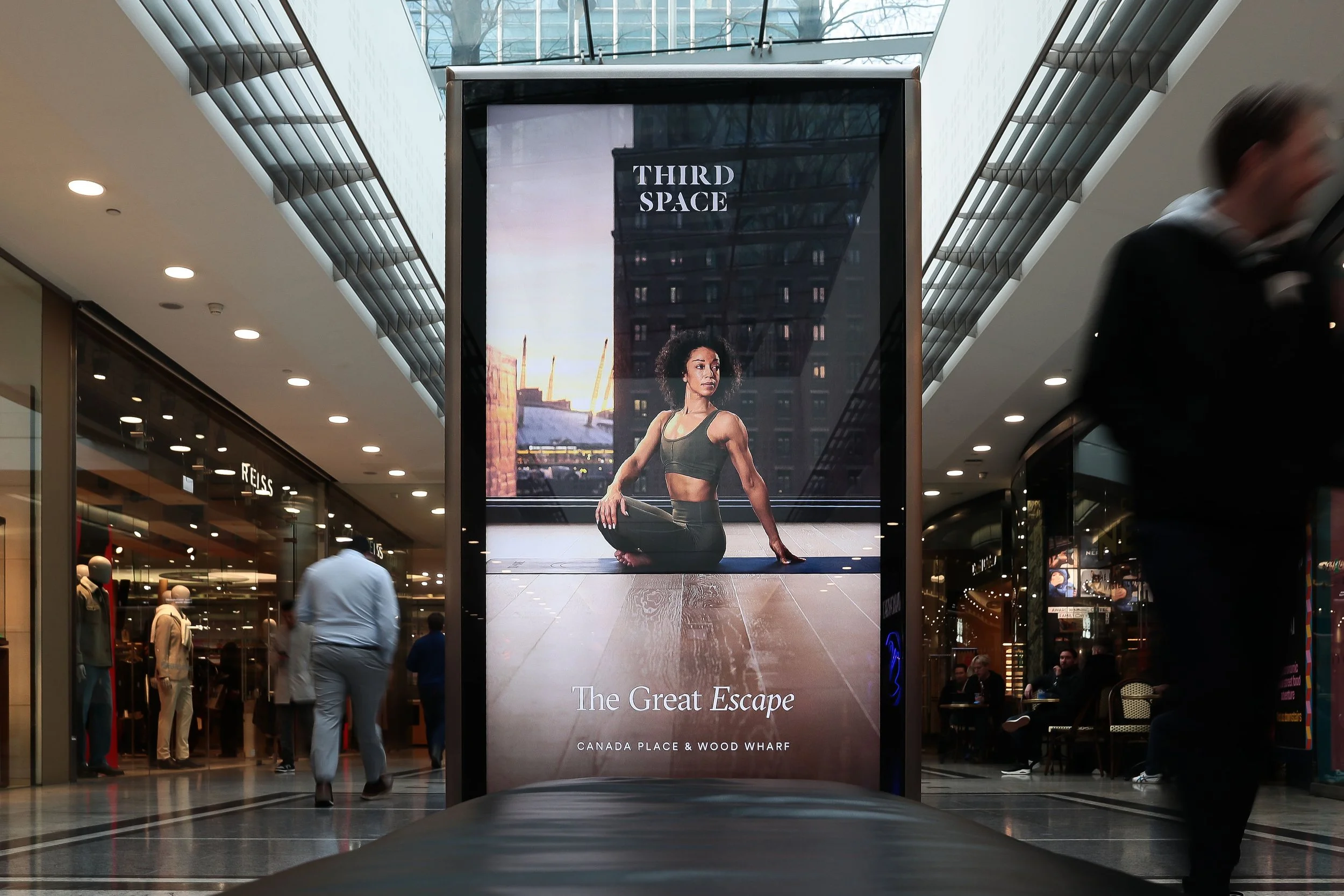 Digital advertisement display in a shopping mall showing a woman in yoga attire in a yoga pose with the text "Third Space" and "The Great Escape," located at Canada Place and Wood Wharf.