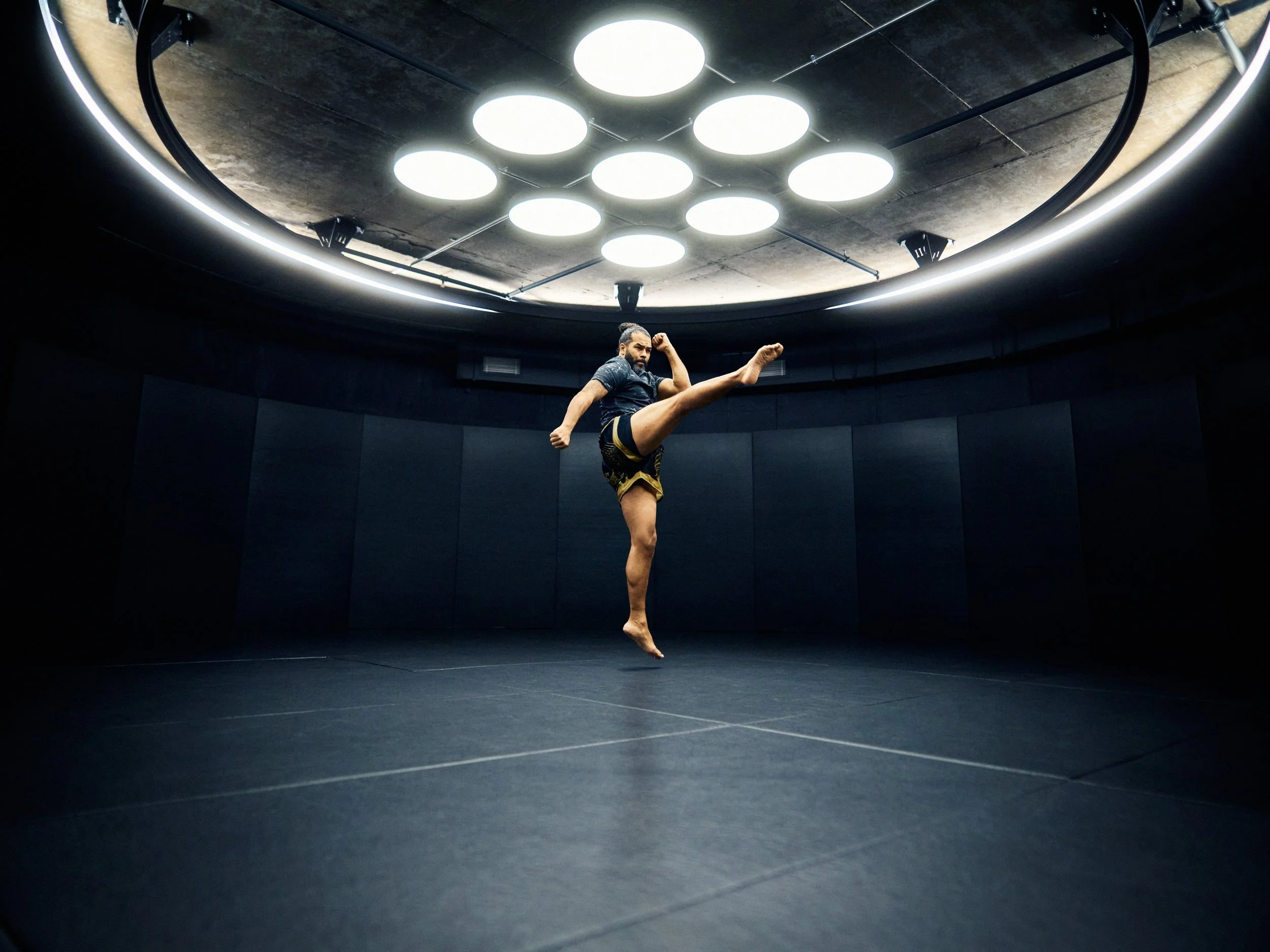A male martial artist practicing a high kick in a dimly lit training room with circular ceiling lights.