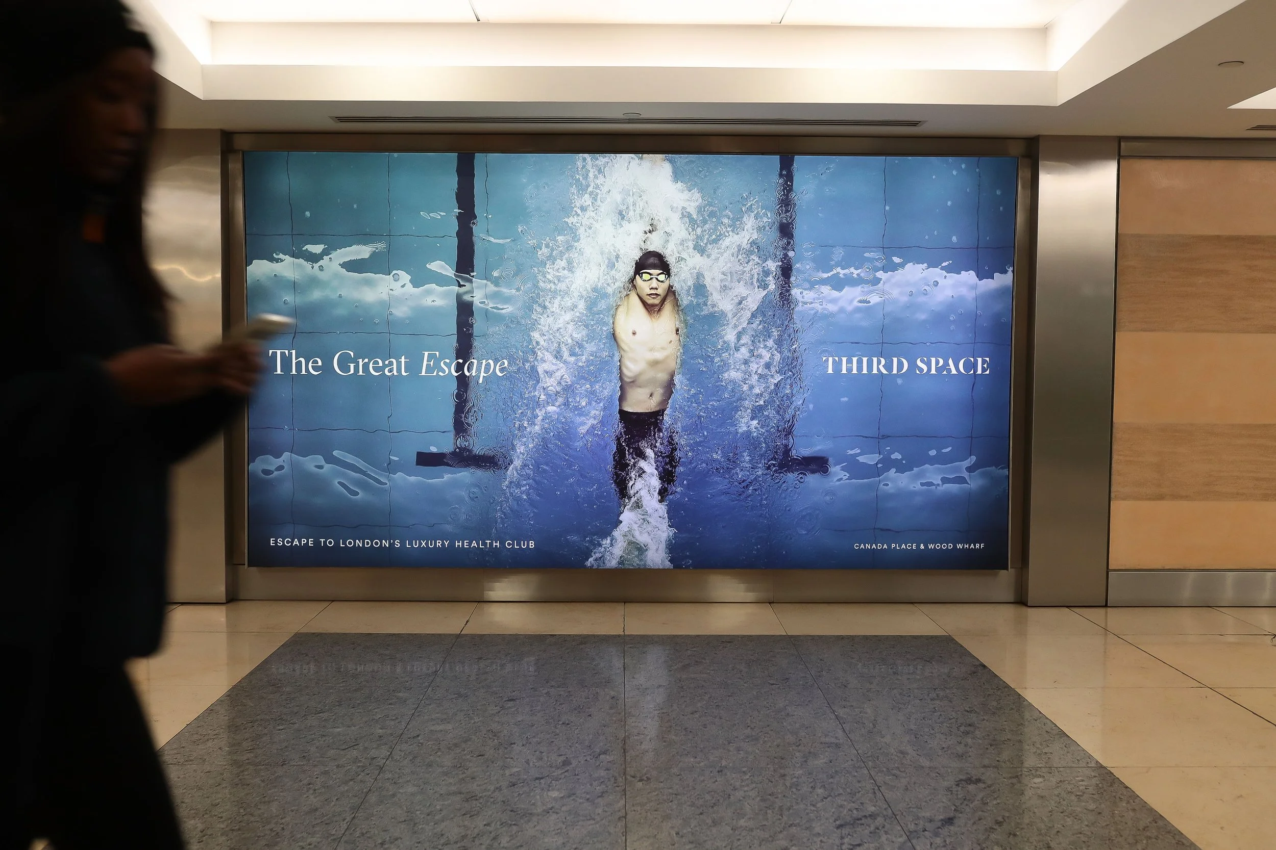 A large advertisement featuring a man swimming underwater with goggles, with text that reads 'The Great Escape' and 'Third Space'. The background resembles a pool with water splashes. The ad is located in an indoor setting, likely a shopping mall or 
