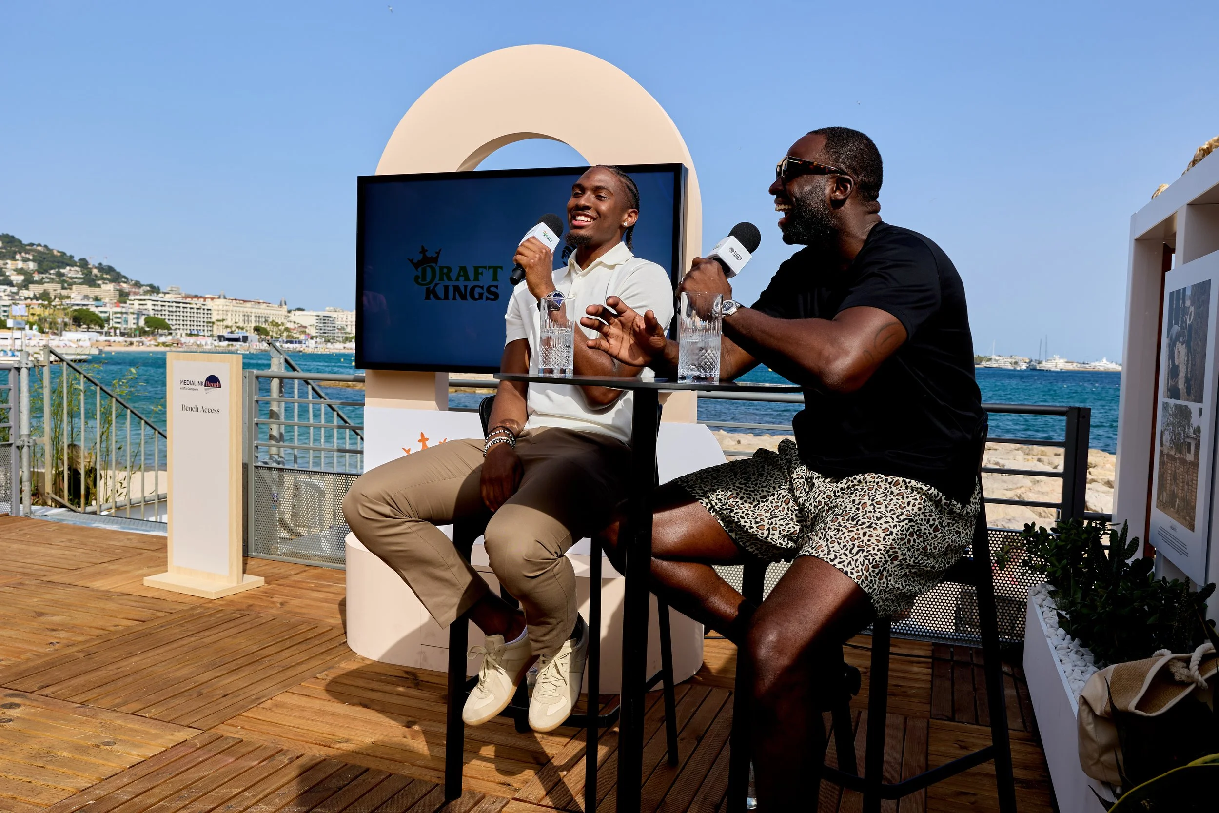 Two men are seated at a table outdoors near the water, holding microphones during a discussion or interview at an event with a screen displaying "Draft Kings" behind them. The setting is sunny with a cityscape and water in the background.