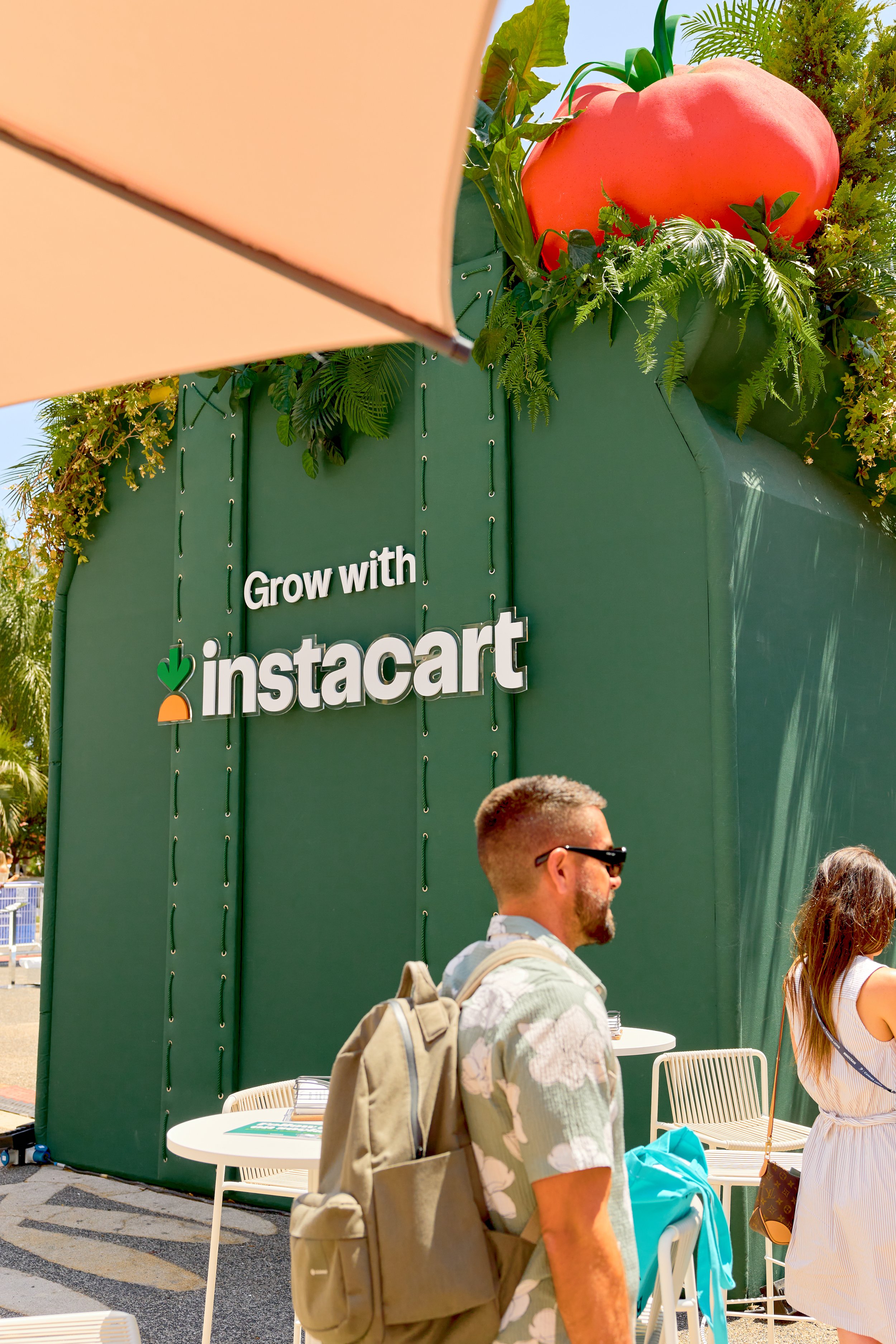 People walking near an outdoor exhibit with a green backdrop featuring the text 'Grow with instacart' and a large tomato sculpture at the top.