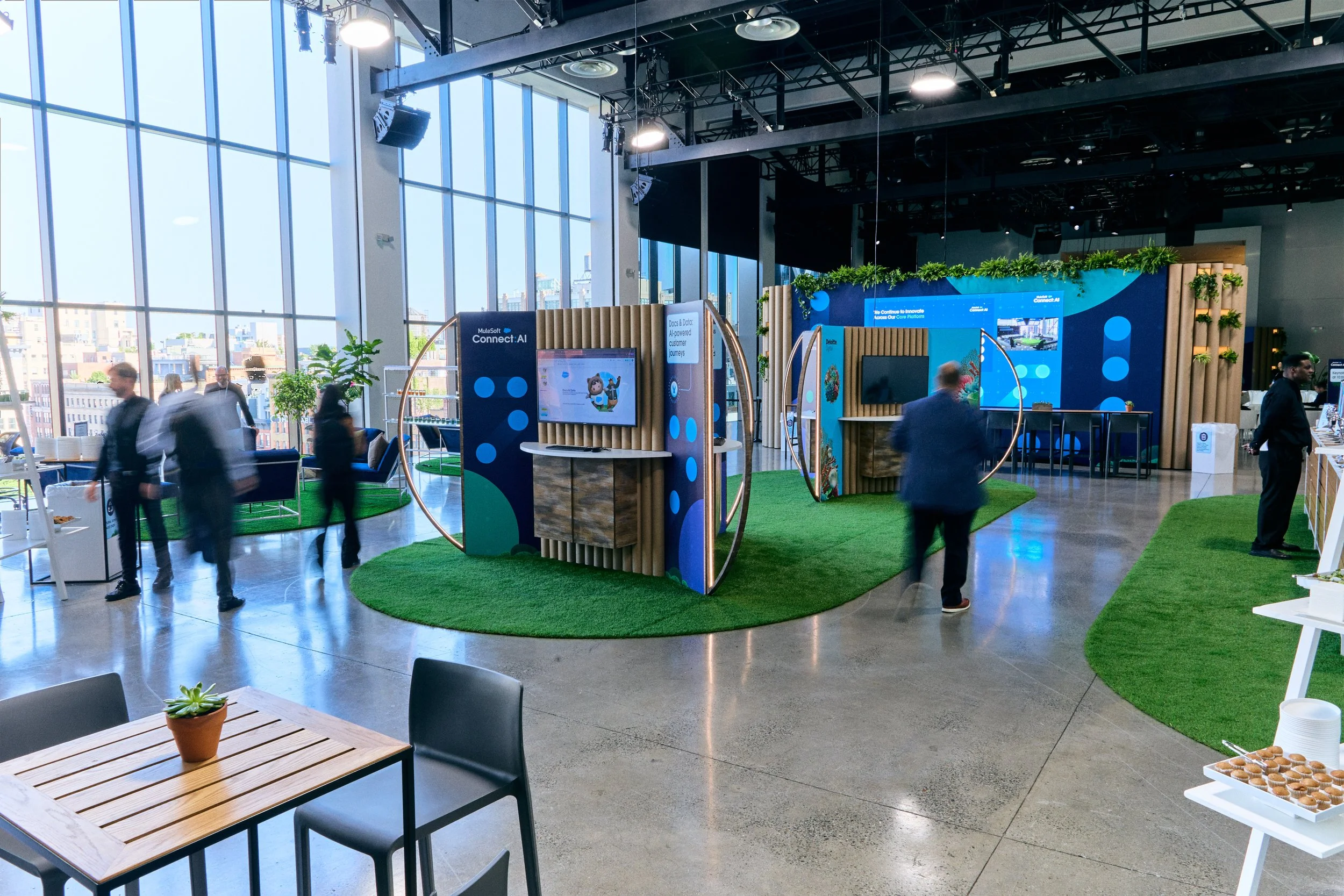 An indoor event space with large glass windows, modern decor, and people walking around. There are display booths with digital screens, a table with snacks, and artificial grass areas for seating.
