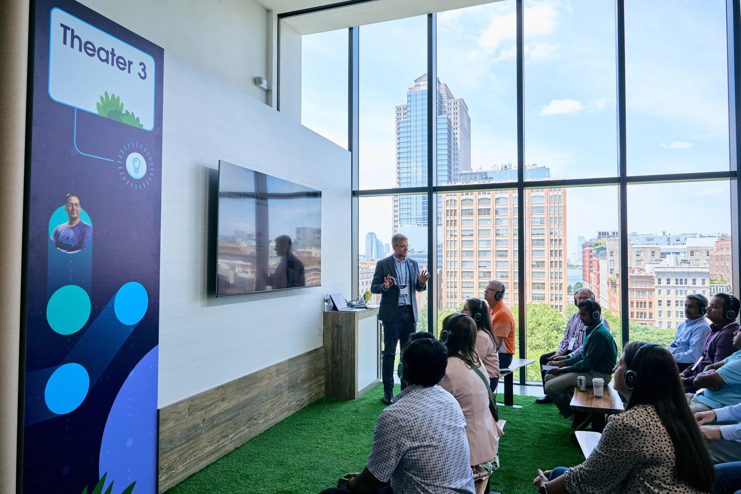 A man in a suit giving a presentation to an audience in a conference room with large windows and a city view, with a standing banner labeled "Theater 3" on the left side.