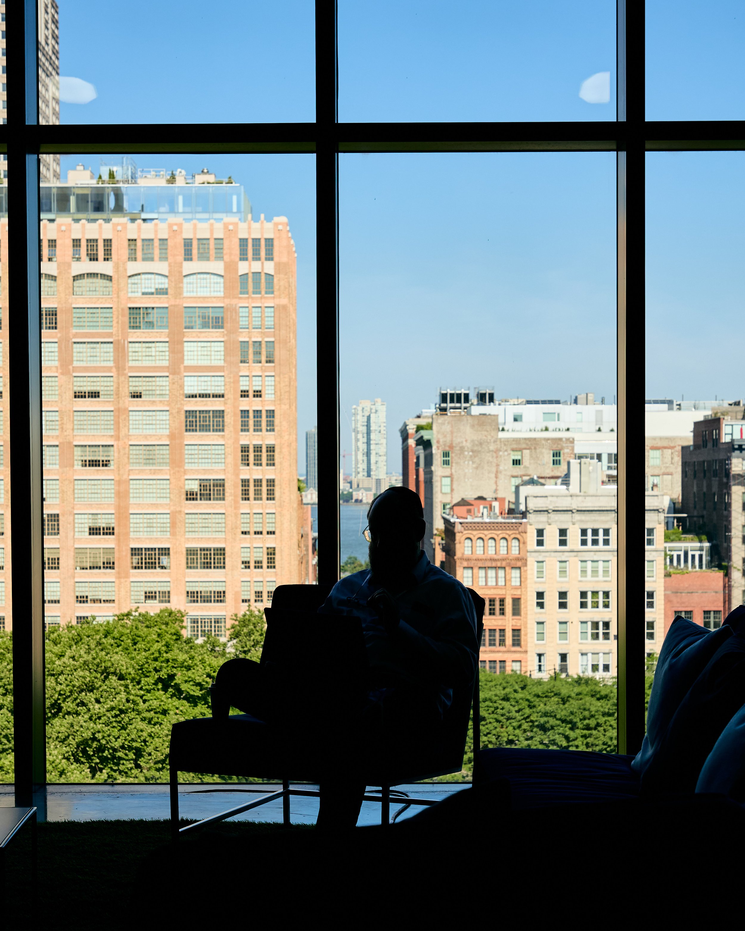 Silhouette of a person sitting in an armchair by a large window, overlooking a cityscape with tall buildings, trees, and a body of water.