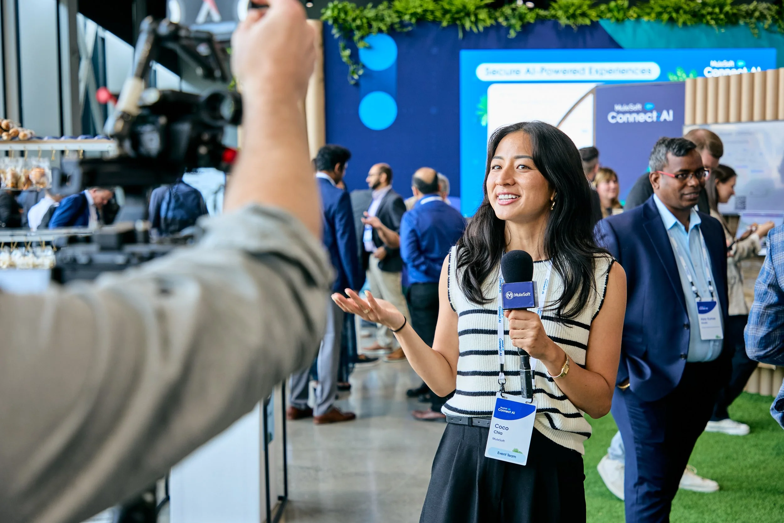A woman with long black hair and a striped sleeveless top holding a microphone, smiling and speaking at a conference, with people and large digital screens in the background.