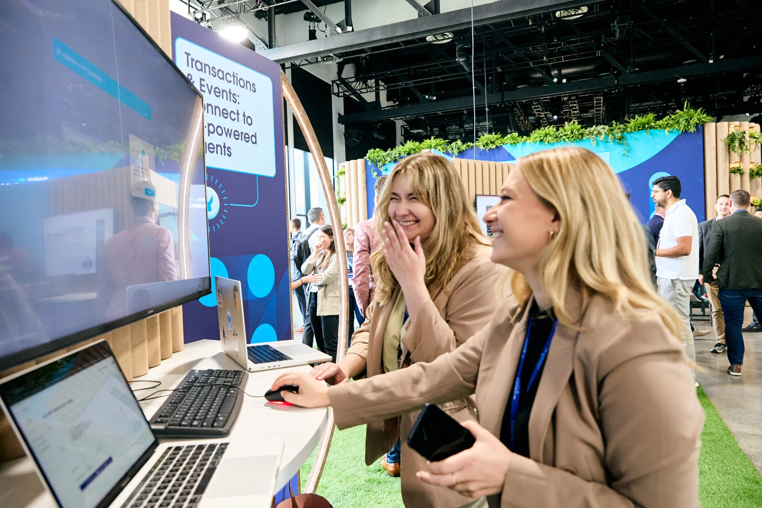 Two women at a trade show or conference, laughing and engaging with a display on computers and a large screen showing a presentation.