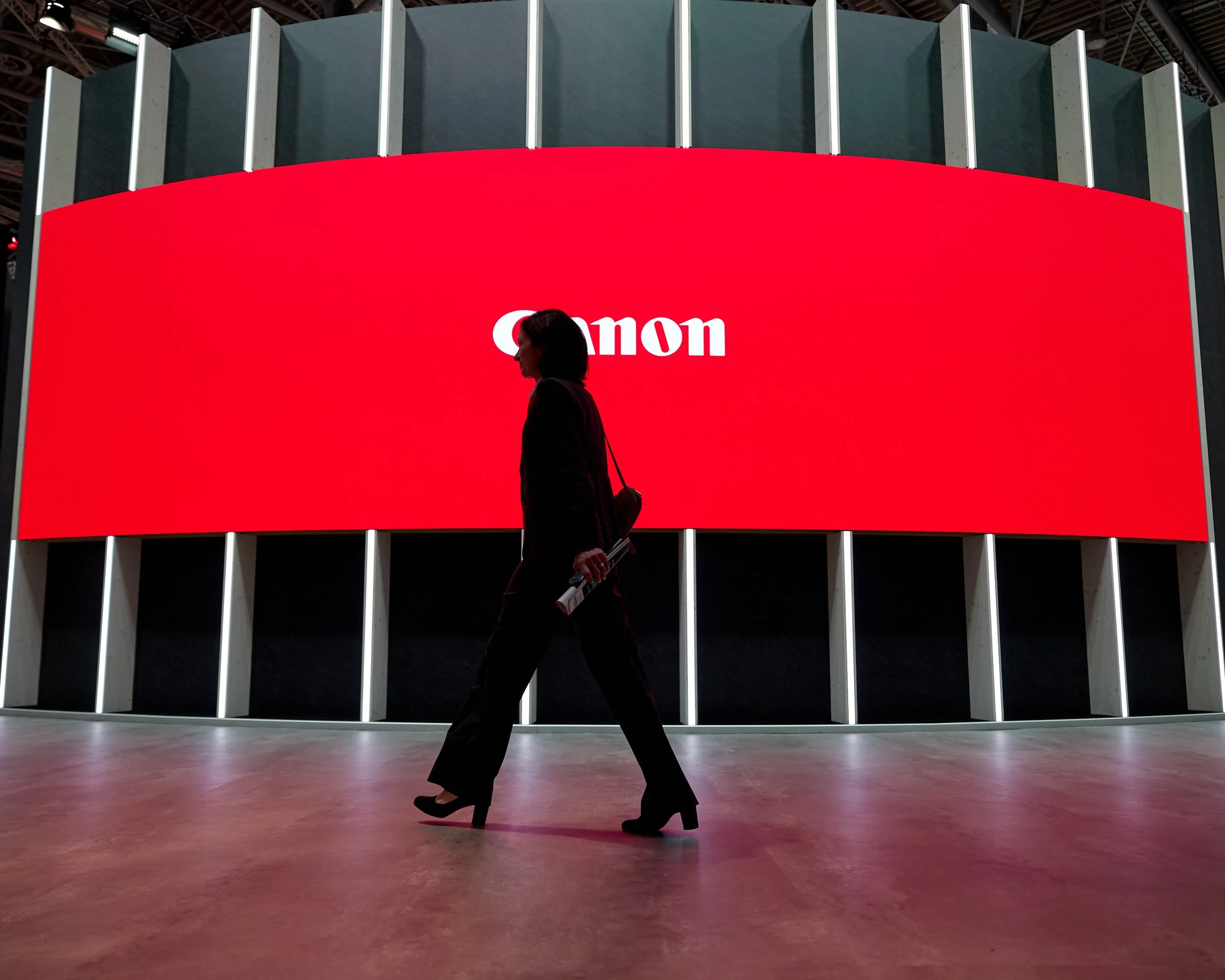 Silhouette of a woman walking in front of a large red digital screen displaying the Canon logo at an exhibition or conference.