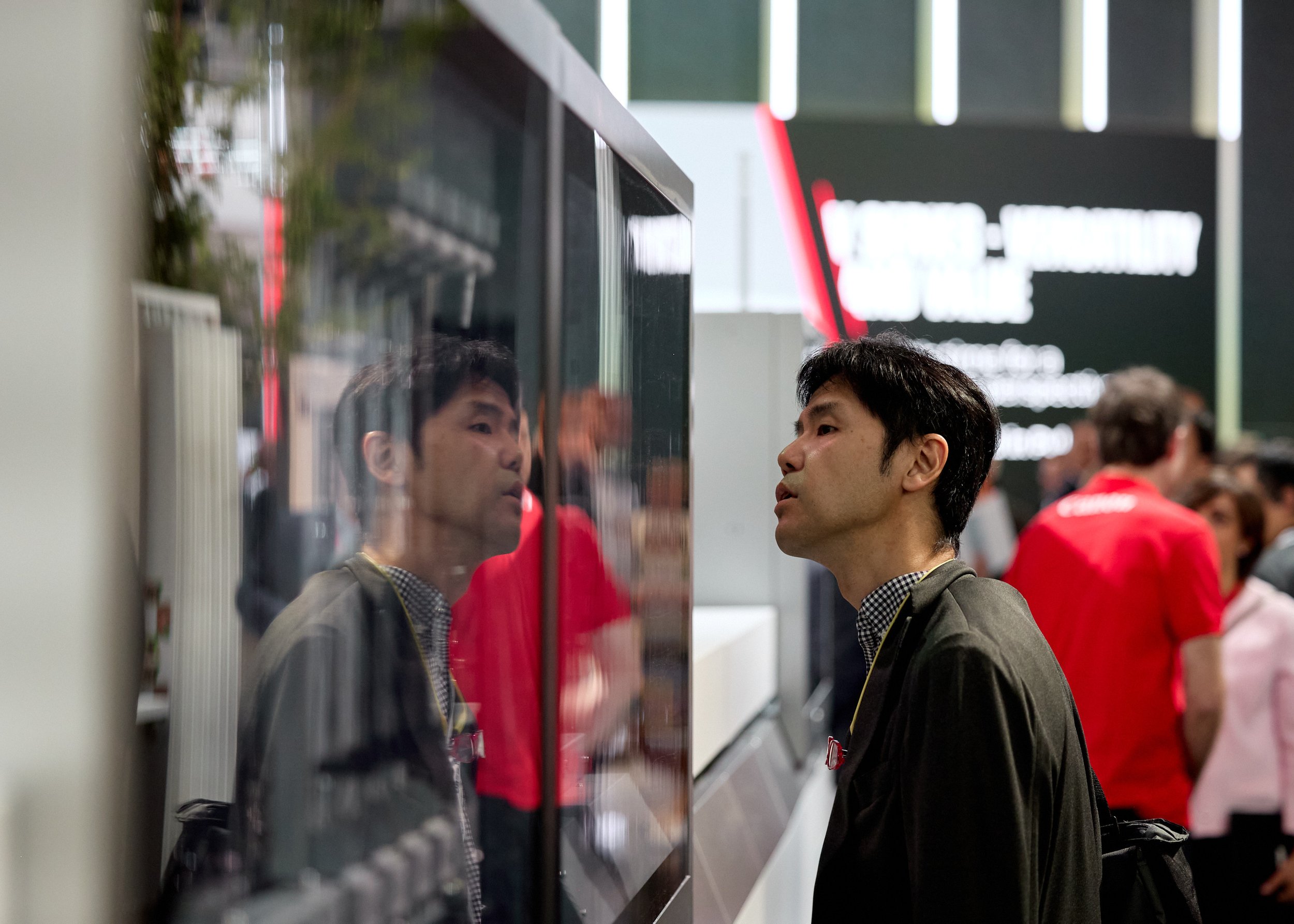 A man with black hair and wearing a dark jacket looks at his reflection in a glass display, with a busy exhibition or trade show background including people and a large screen.