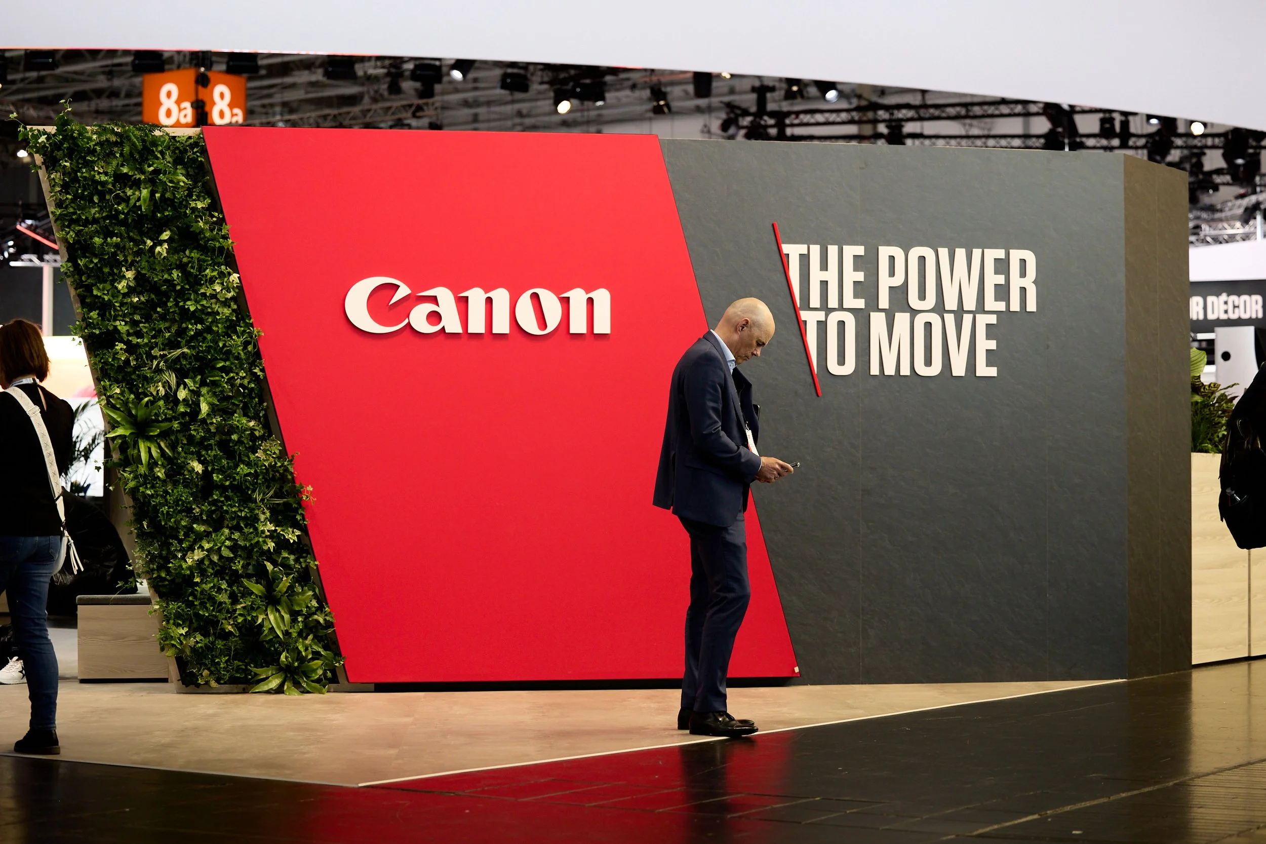 A man in a suit stands on a stage with a large display behind him showing the Canon logo and the slogan "The Power to Move" at a trade show or exhibition.