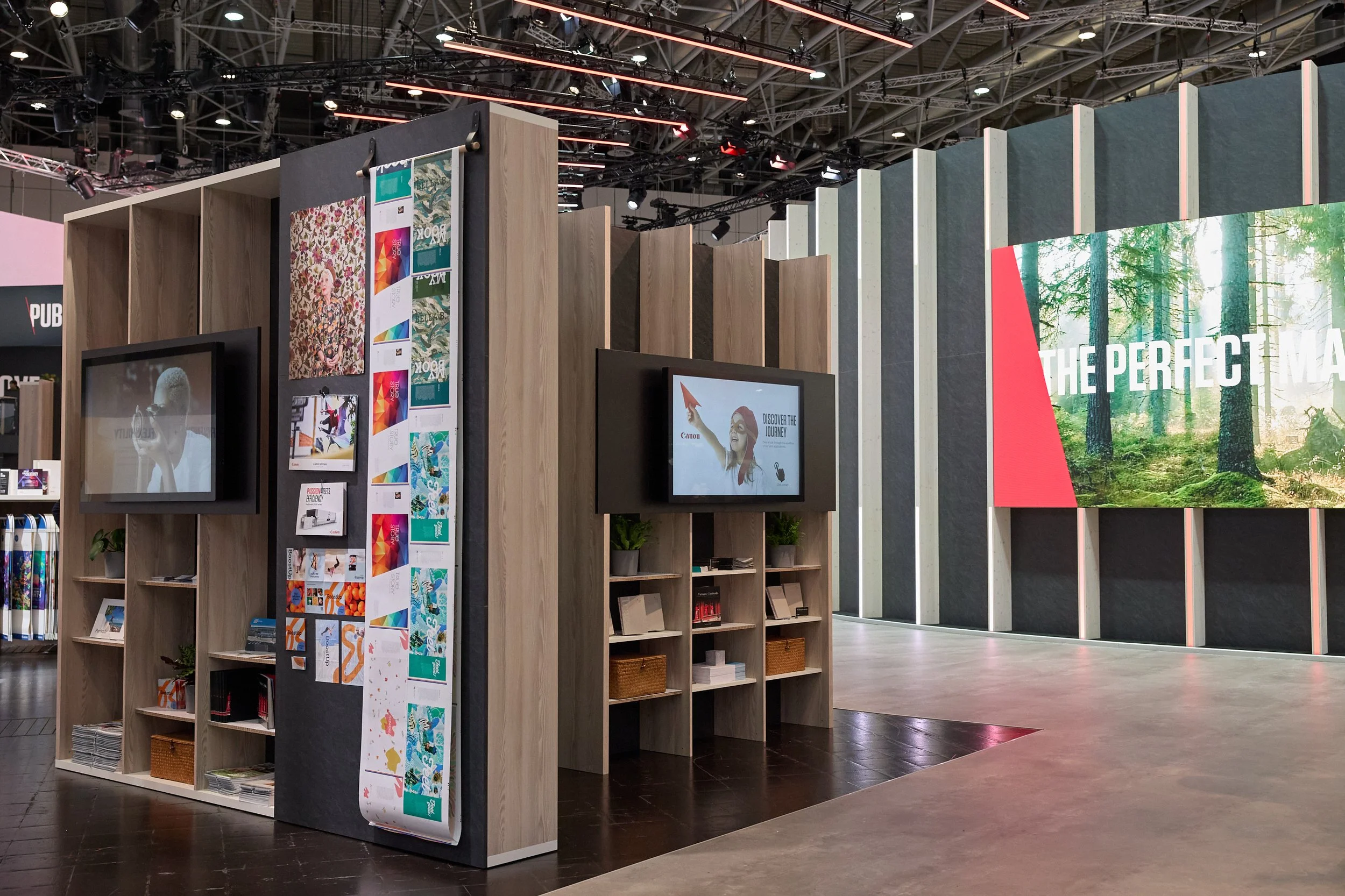 An exhibition booth displaying various wall panels, promotional banners, and two mounted screens showing advertising content, at a trade show or convention center.