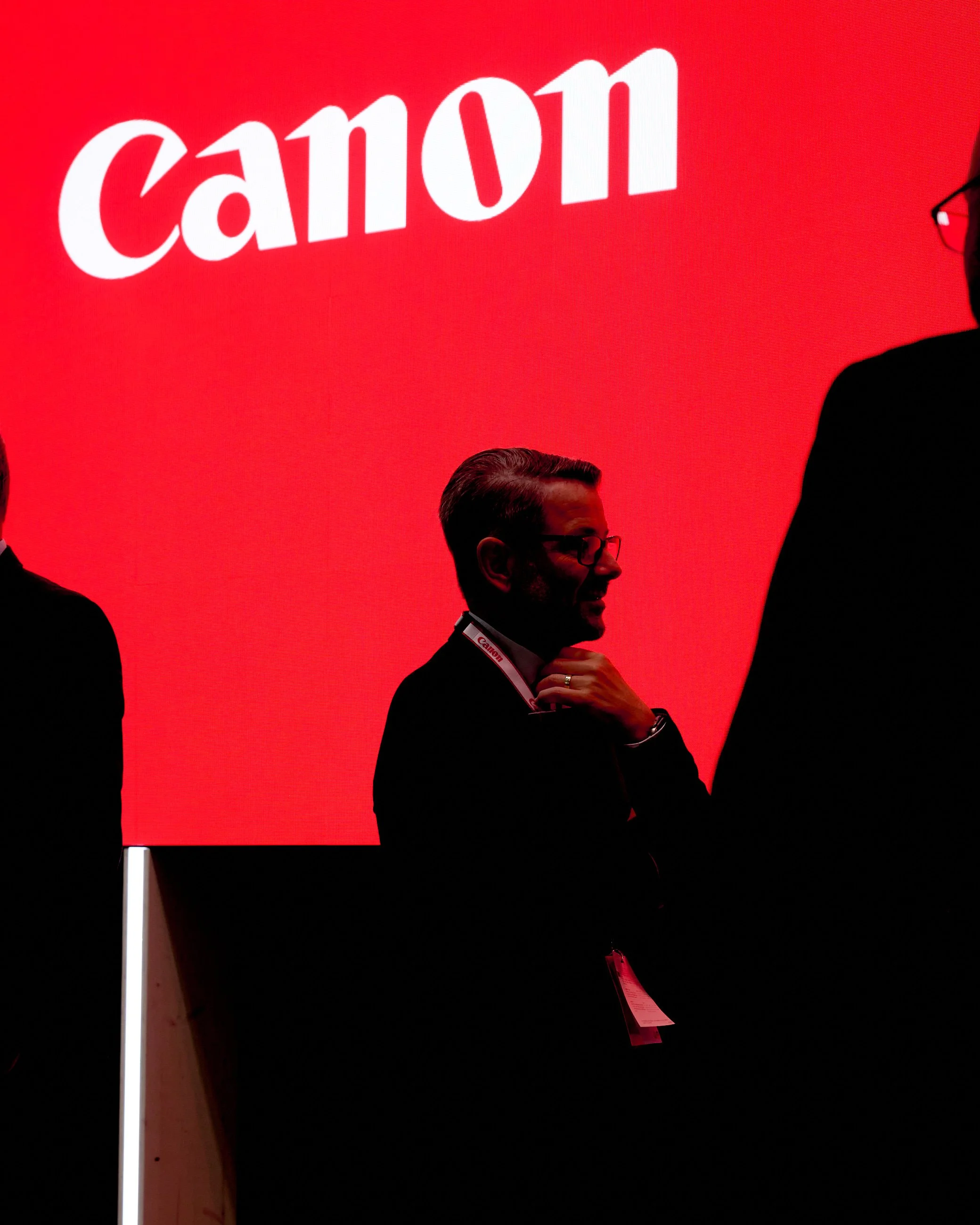 Silhouette of a man with eyeglasses and a beard, standing against a large red backdrop with the white 'Canon' logo, at a professional event or conference.
