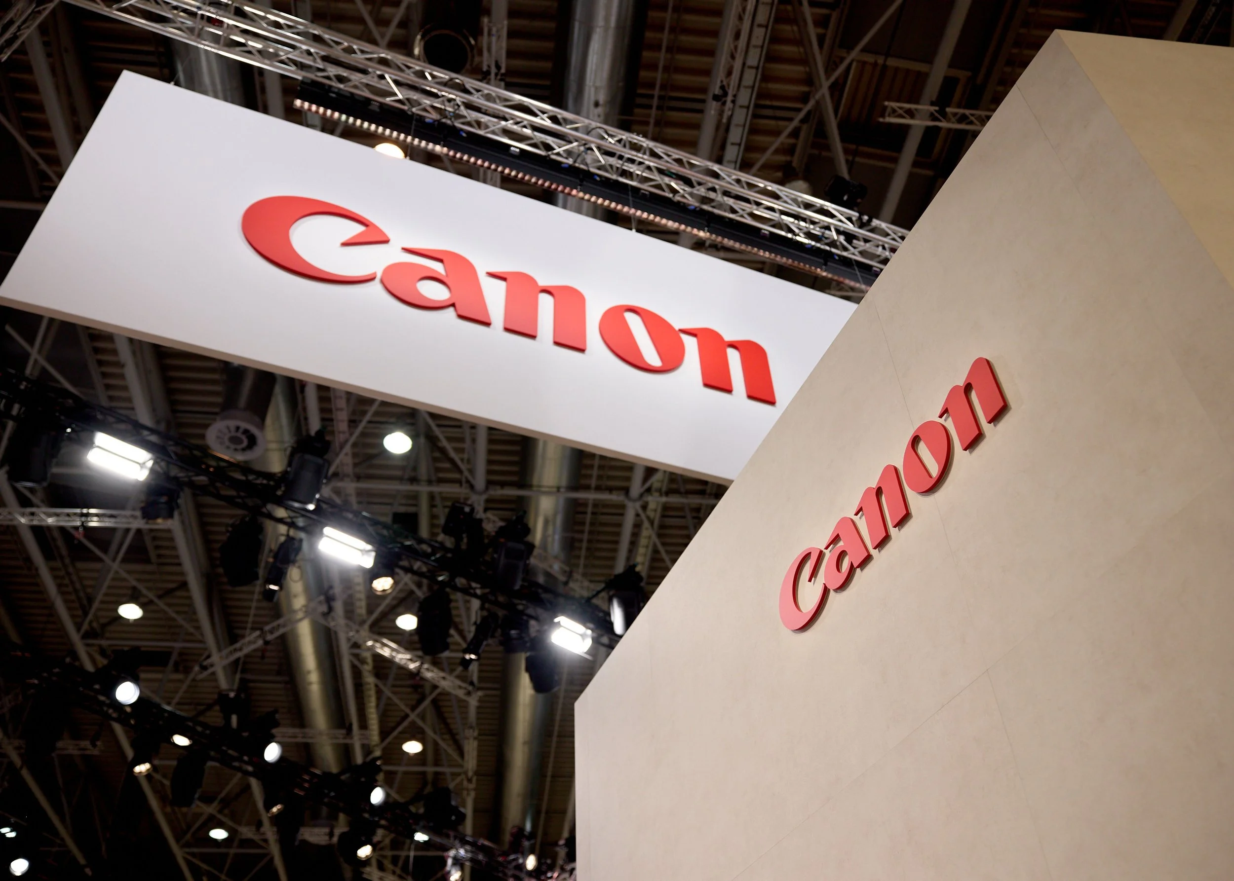 Canon logo displayed on a large overhead sign and a wall at an indoor trade show or convention.