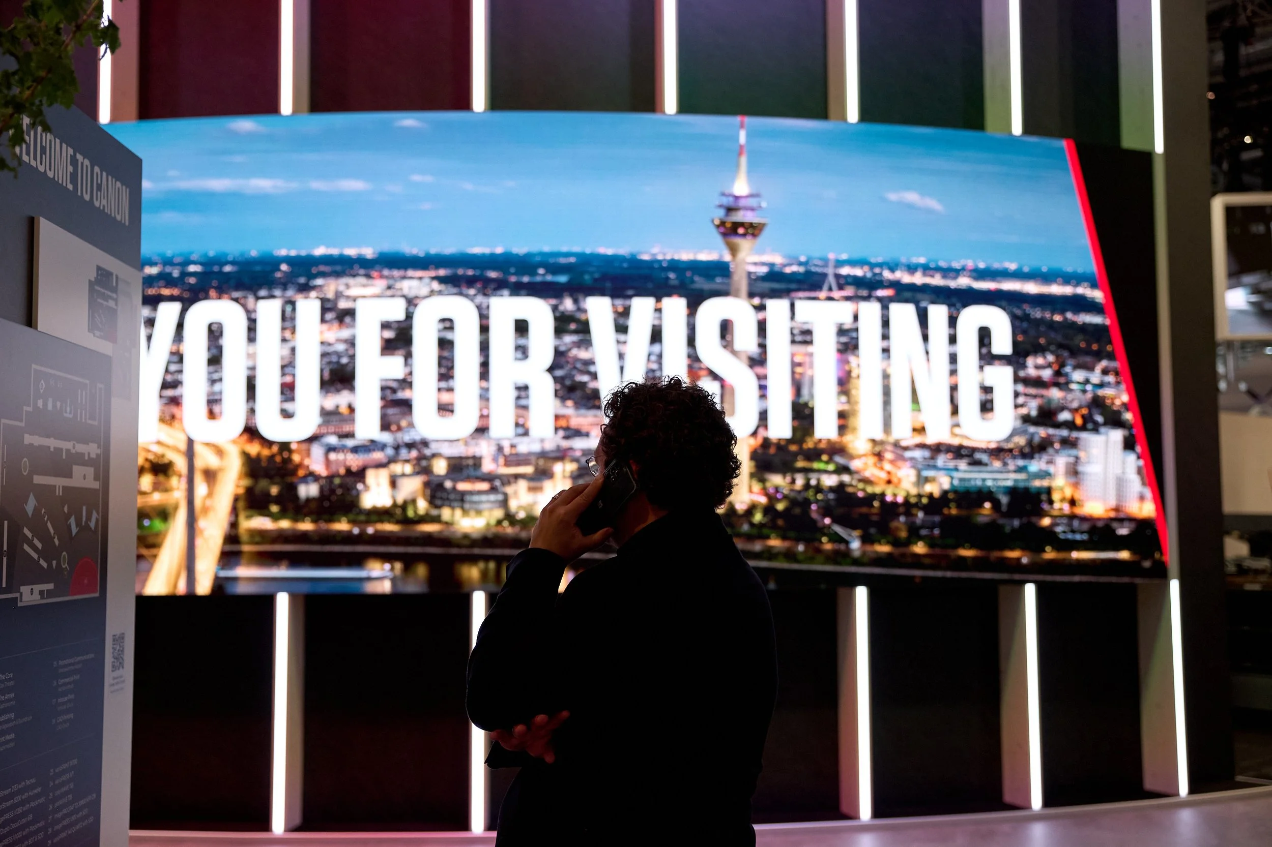 A person talking on a cellphone in front of a large screen that displays the city skyline and the text 'YOU FOR VISITING'.
