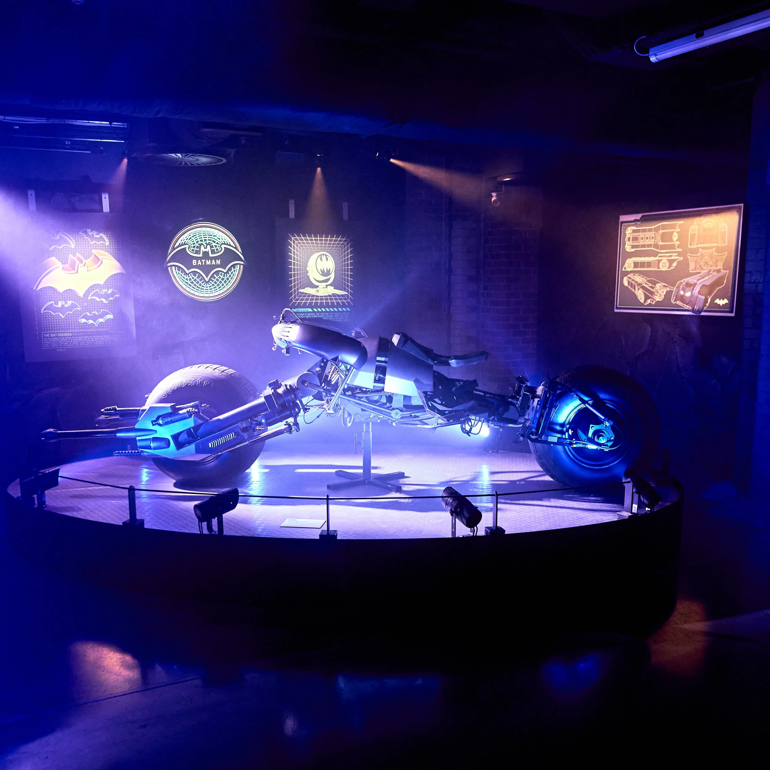 A futuristic Batmobile car on display with neon Batman-themed posters and diagrams on the wall behind it, illuminated by blue and purple lighting.