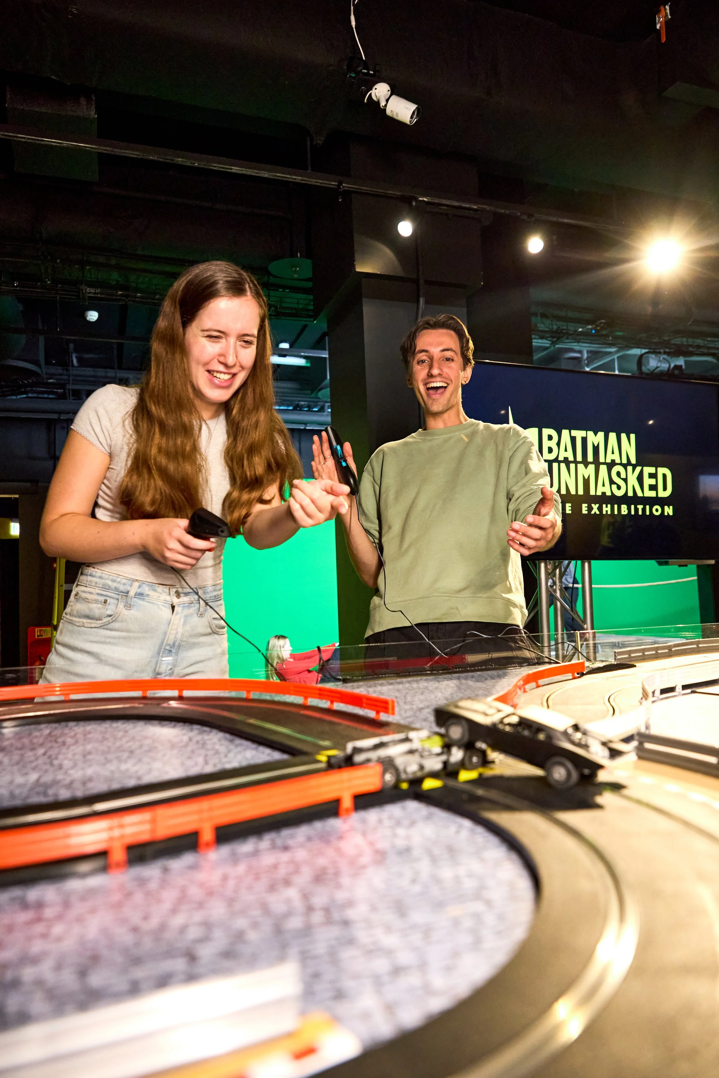 Two young adults, a woman with long wavy hair and a man, are playing with a slot car racing track at an exhibition titled 'Batman Unmasked.' They are smiling and enjoying themselves.