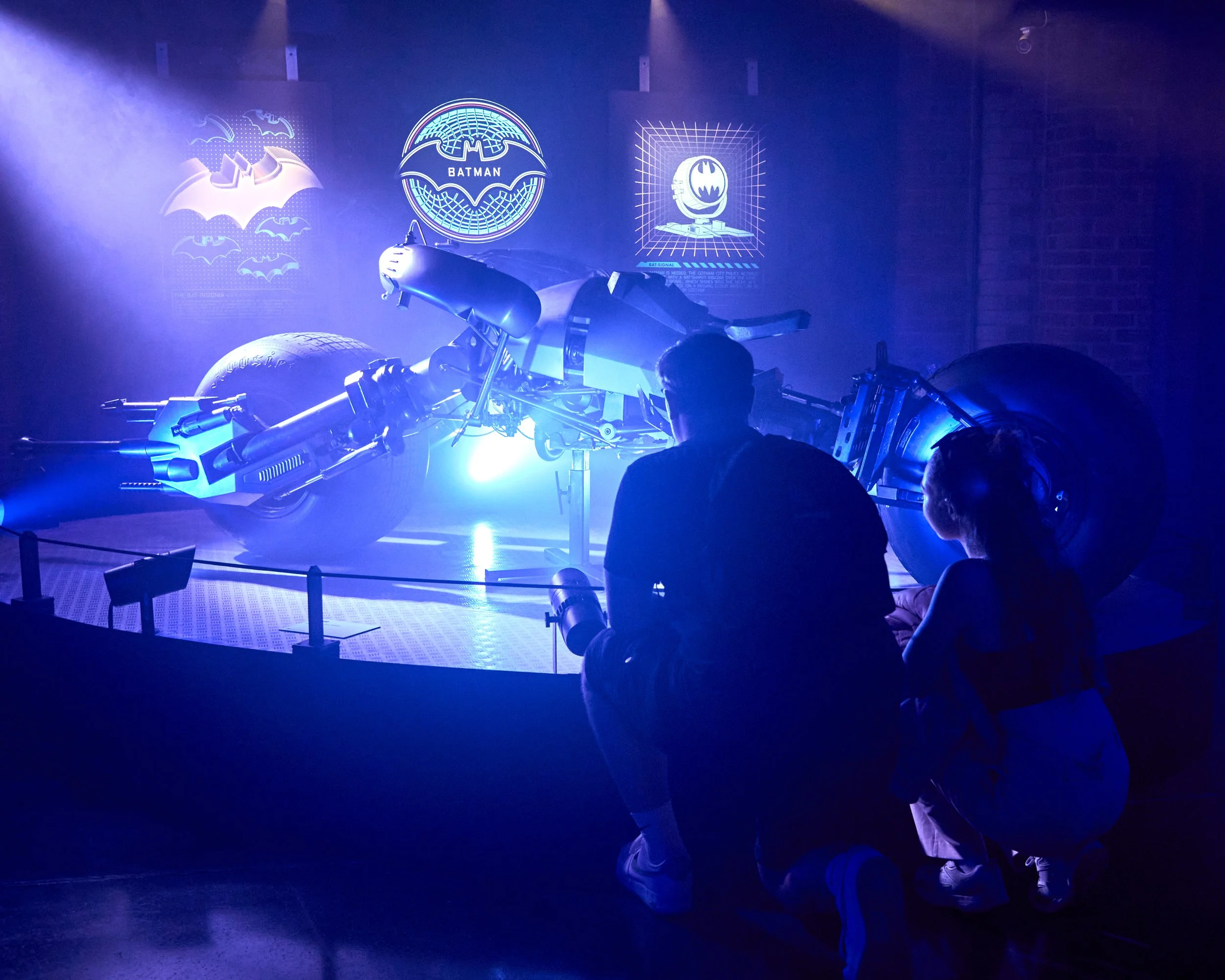 Two people, a man and a woman, kneeling in front of a Batmobile from the Batman franchise, displayed behind a protective barrier in a museum or exhibit, with Batman logos and art on the wall in blue lighting.