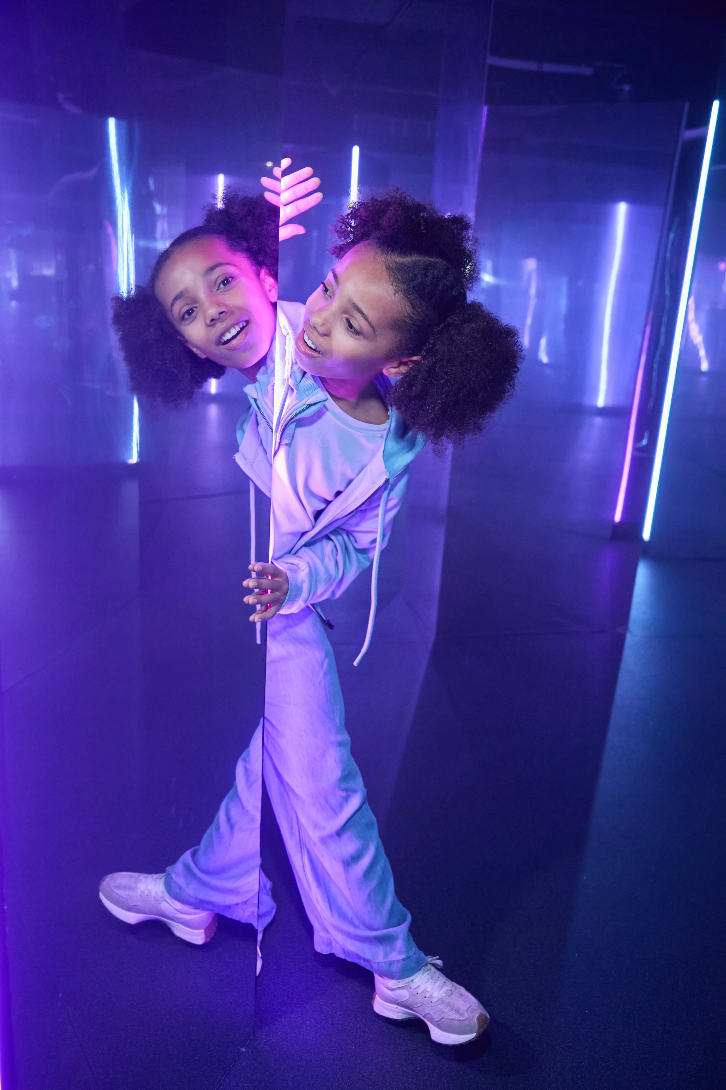 A young girl with curly hair in a colorful, illuminated room with purple and blue lighting, peeking through a mirror or panel.