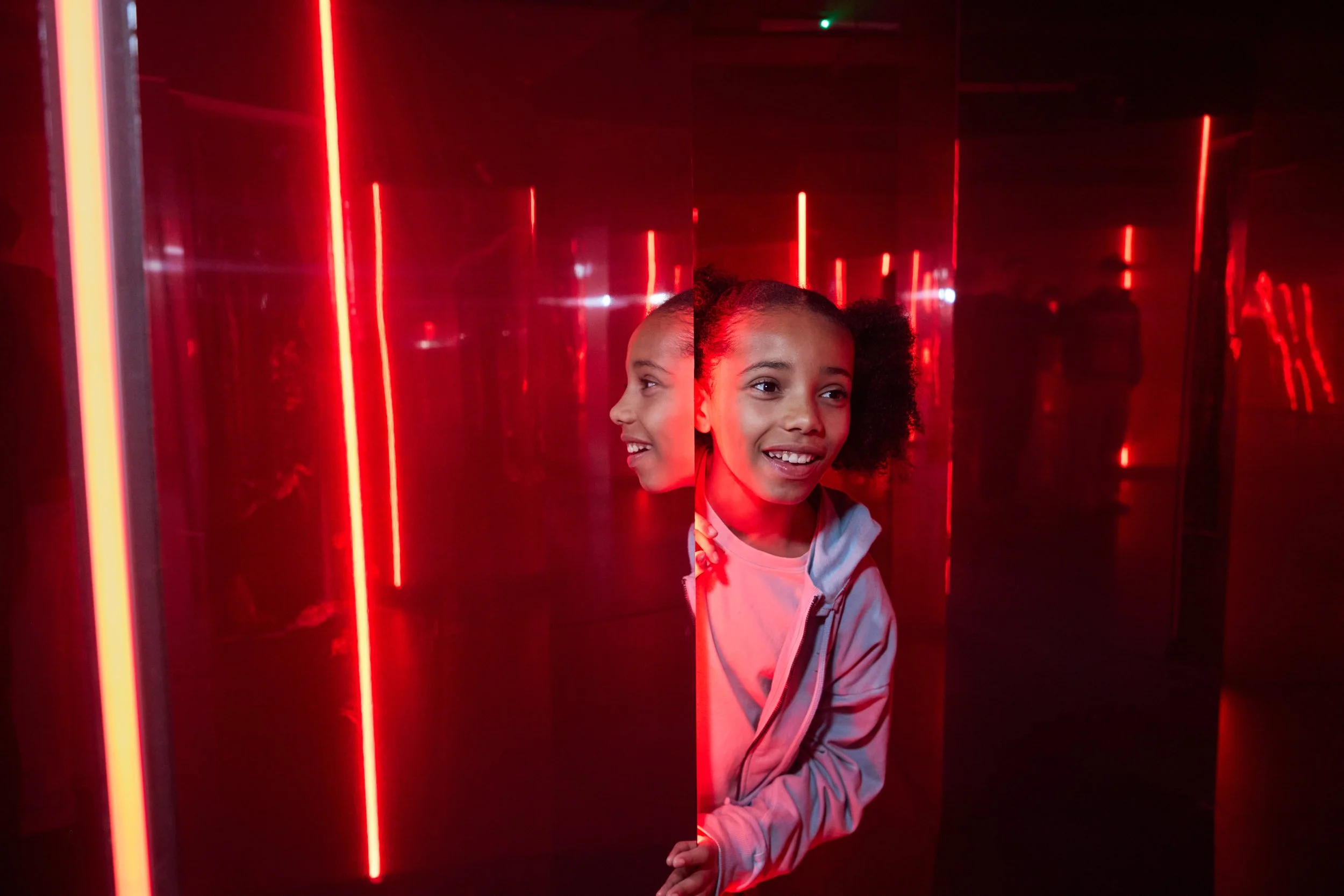A young girl with curly hair peeks out from behind a mirror in a room with red neon lights.