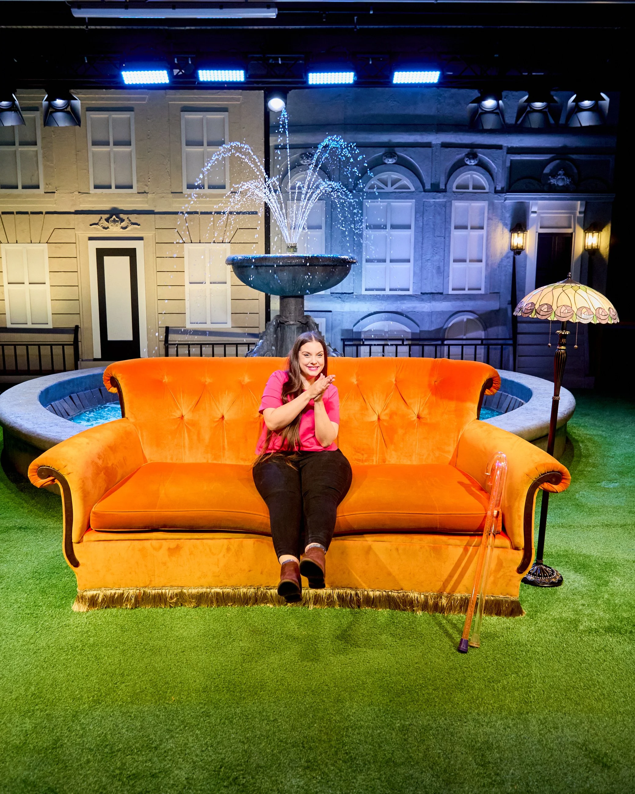 A woman in a pink shirt and dark pants sitting on an orange vintage sofa on a stage set designed to look like a park with a fountain and a backdrop of a city street with buildings, windows, and street lamps. She is smiling and clasping her hands.
