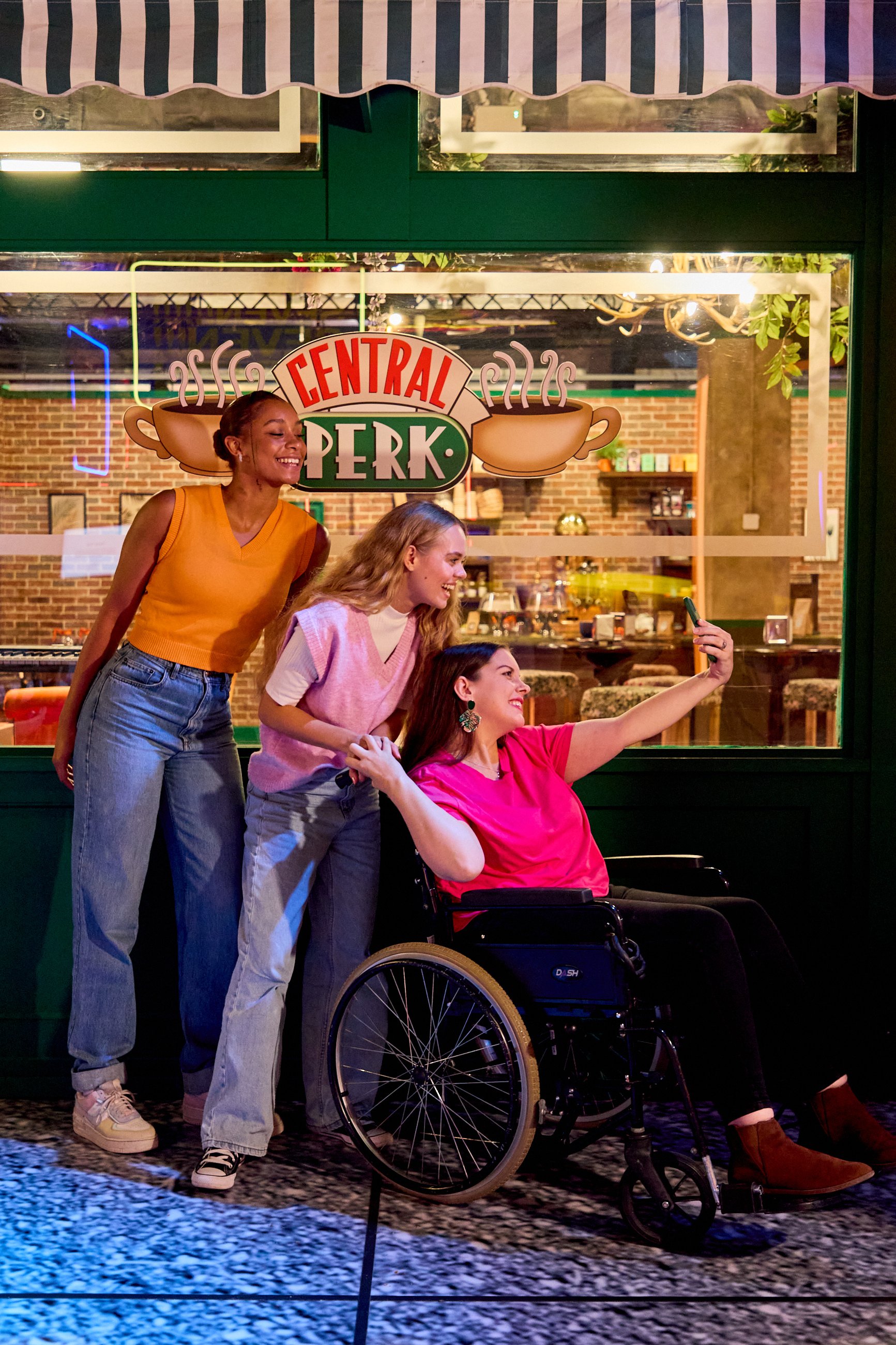 Three women taking a selfie in front of a restaurant window with a 'Central Perk' sign, one woman in a wheelchair, all smiling and enjoying their time.
