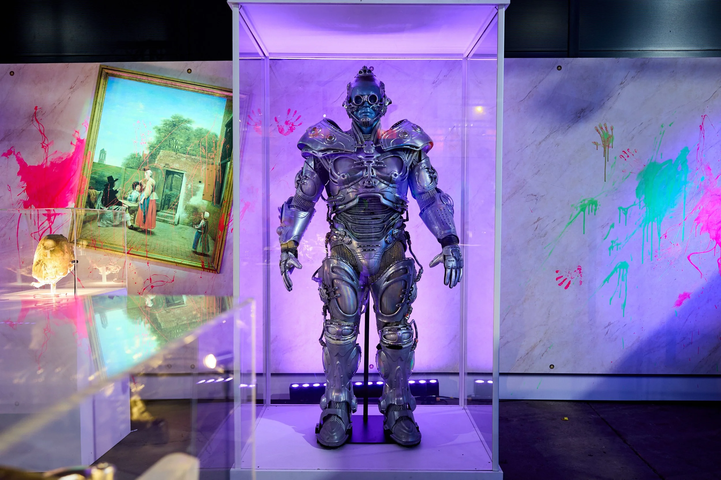A robotic humanoid figure displayed in a glass case with purple lighting, surrounded by colorful splashes of paint on the wall and a painting in the background.