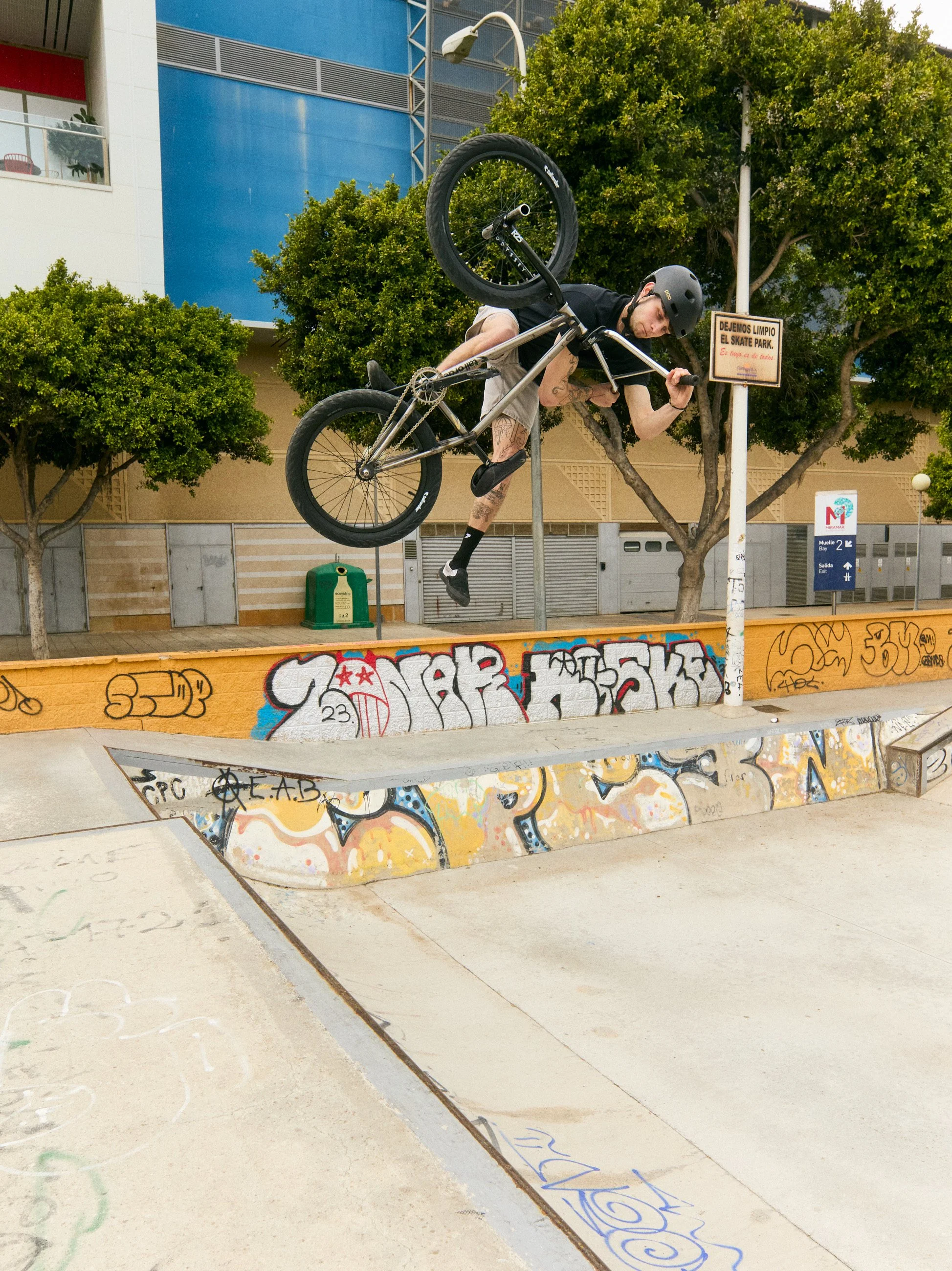 A young BMX rider performing a trick at a skate park, mid-air on his bike, wearing a helmet and black clothing, with graffiti on the ramps and a city building in the background.