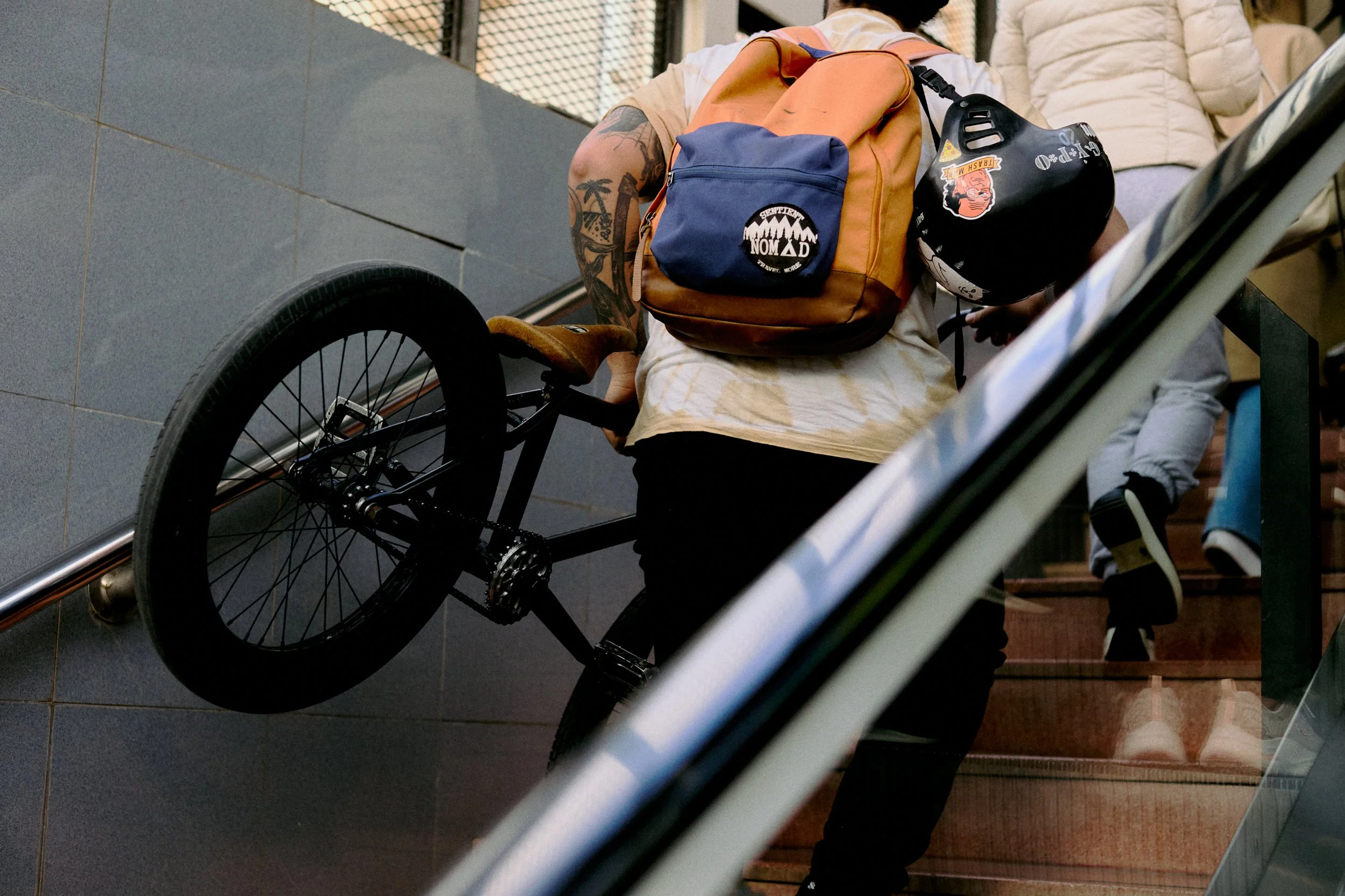 Person with tattoos riding a black BMX bike down the steps of an indoor staircase, wearing a beige shirt, orange and blue backpack, and some stickers on their helmet.
