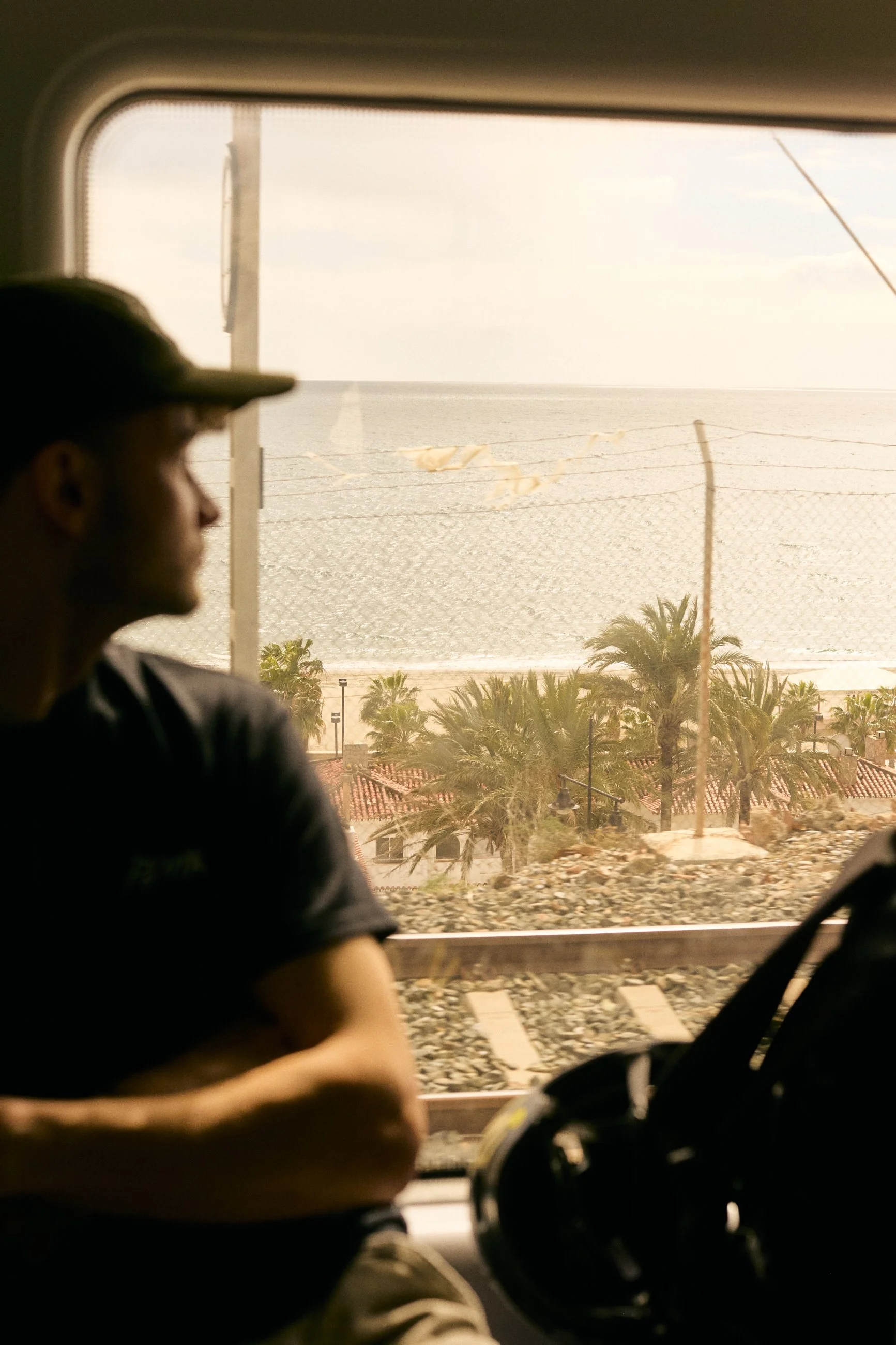 A person sitting inside a vehicle, looking out the window at a coastal scene with palm trees, a fence, and the ocean in the background.
