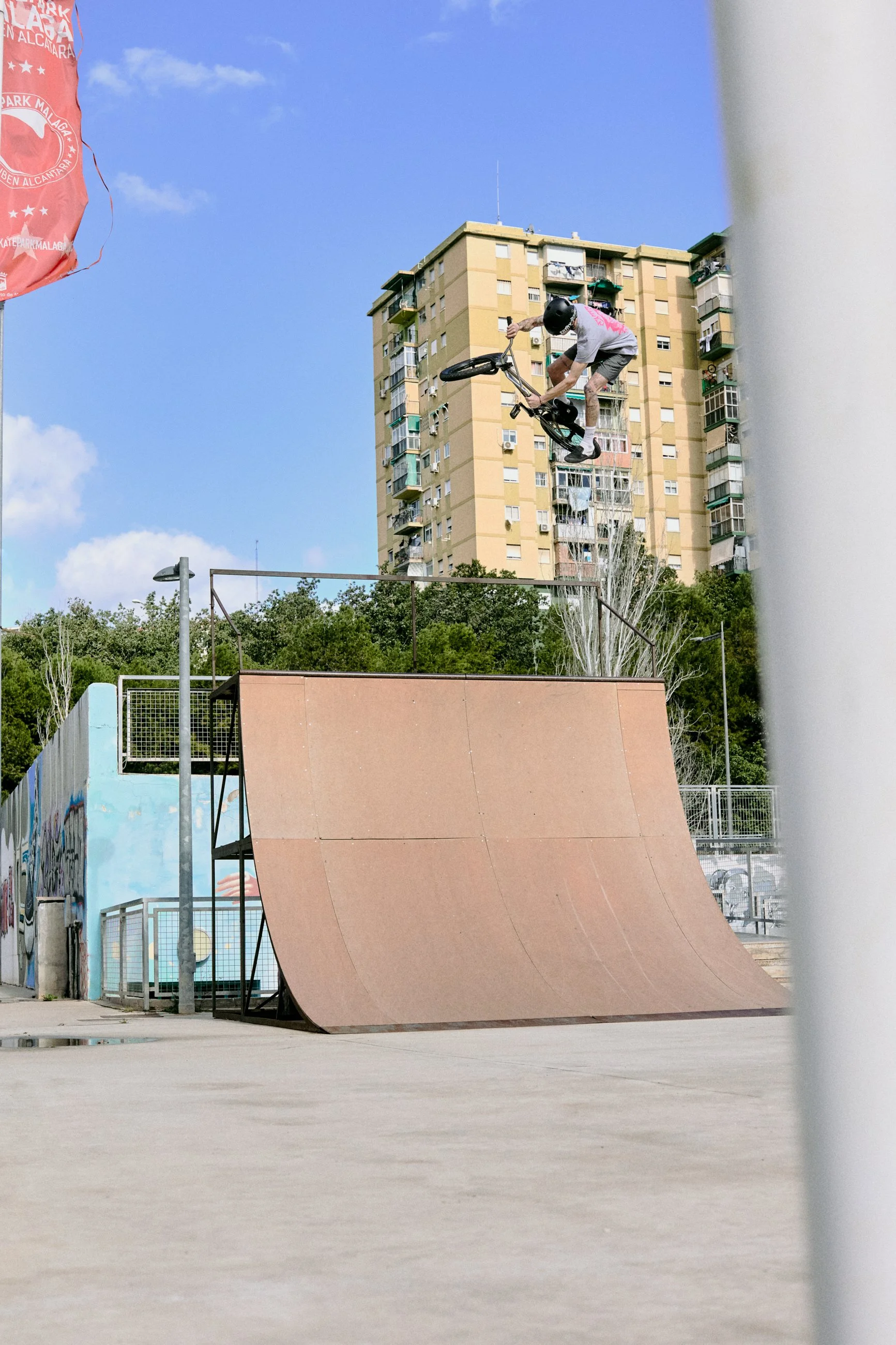 A person performing a BMX trick in mid-air at a skatepark, with a high-rise building and trees in the background.