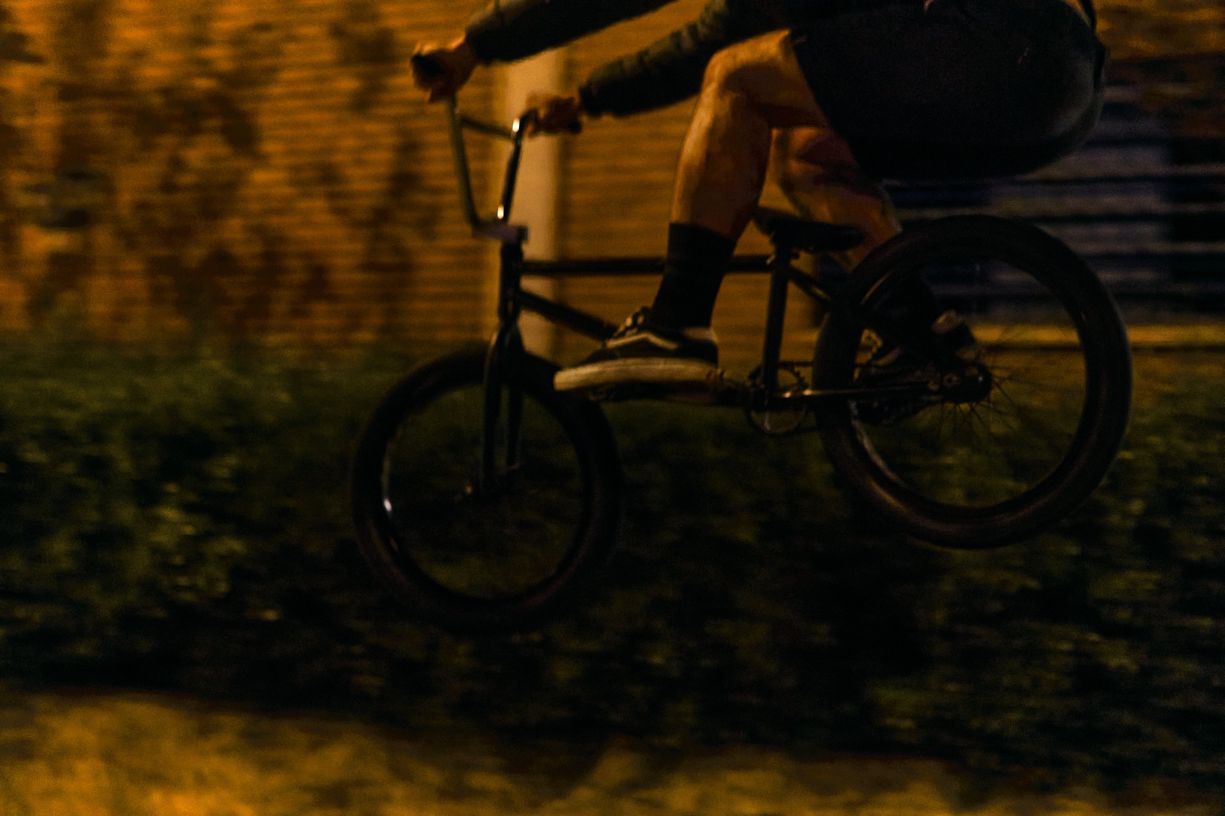 Person riding a bicycle at night on a dark street with brick wall and greenery in the background.