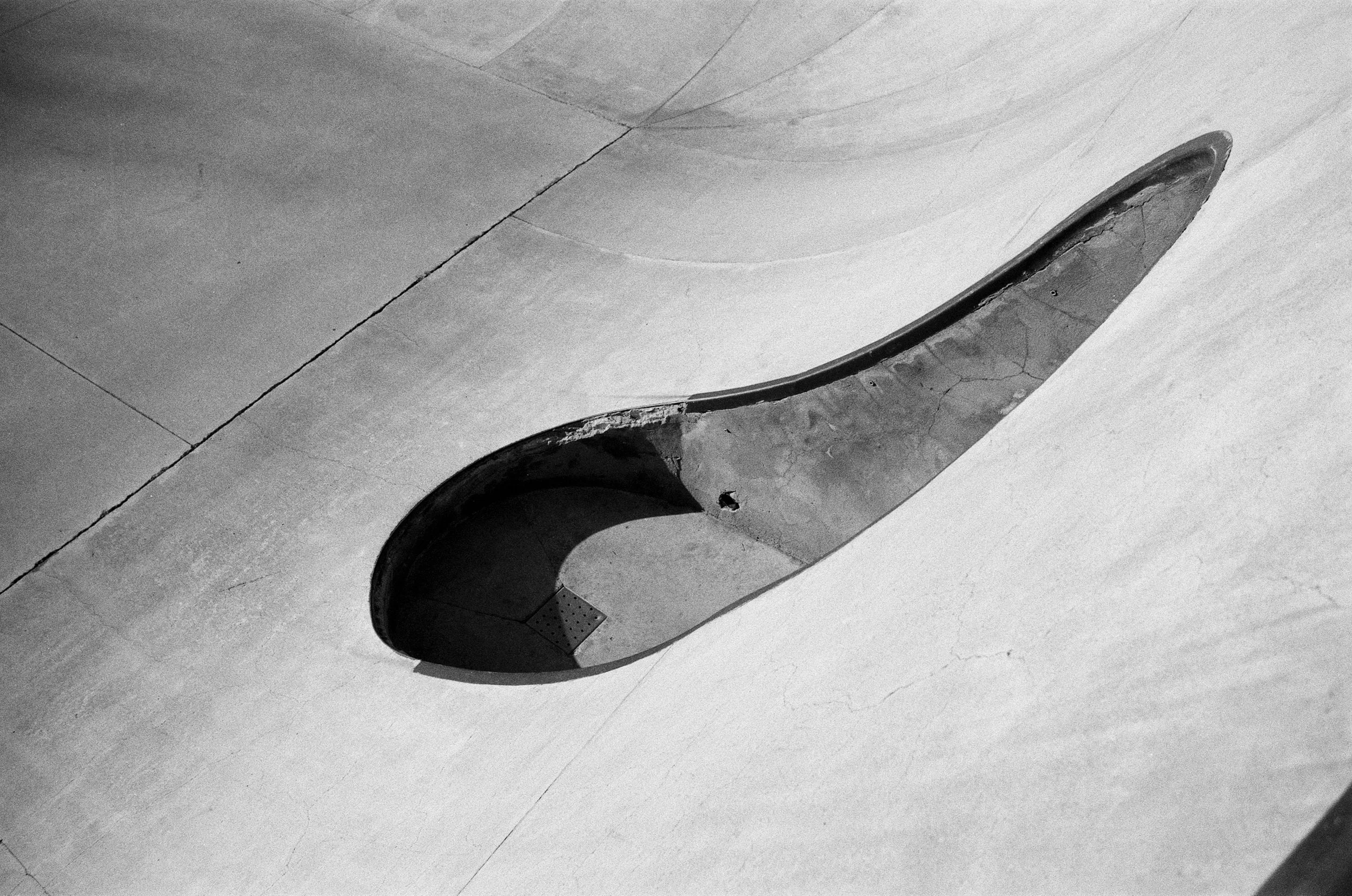 A black and white photograph of a partially buried, curved, and elongated concrete structure resembling a spillway or drainage channel within a concrete floor.