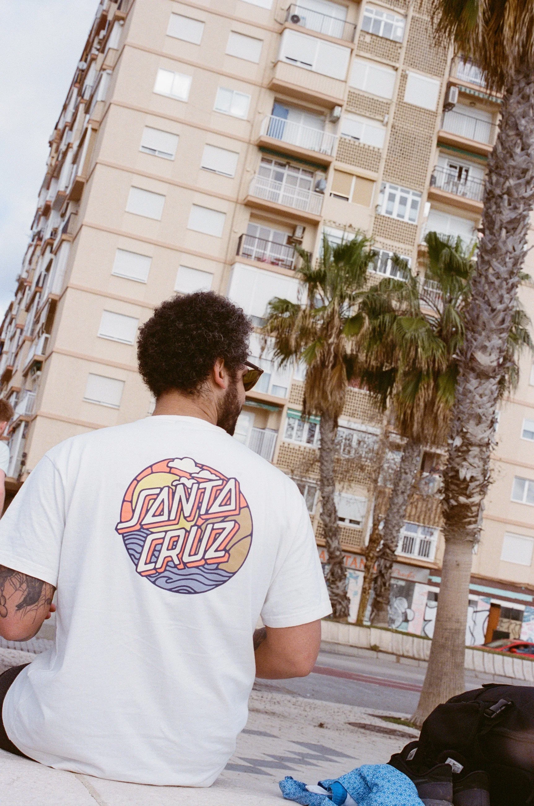 Back of a man with curly hair, wearing sunglasses and a white T-shirt that says 'Santa Cruz' with a sunset, waves, and mountains, sitting outdoors near palm trees with apartment buildings in the background.