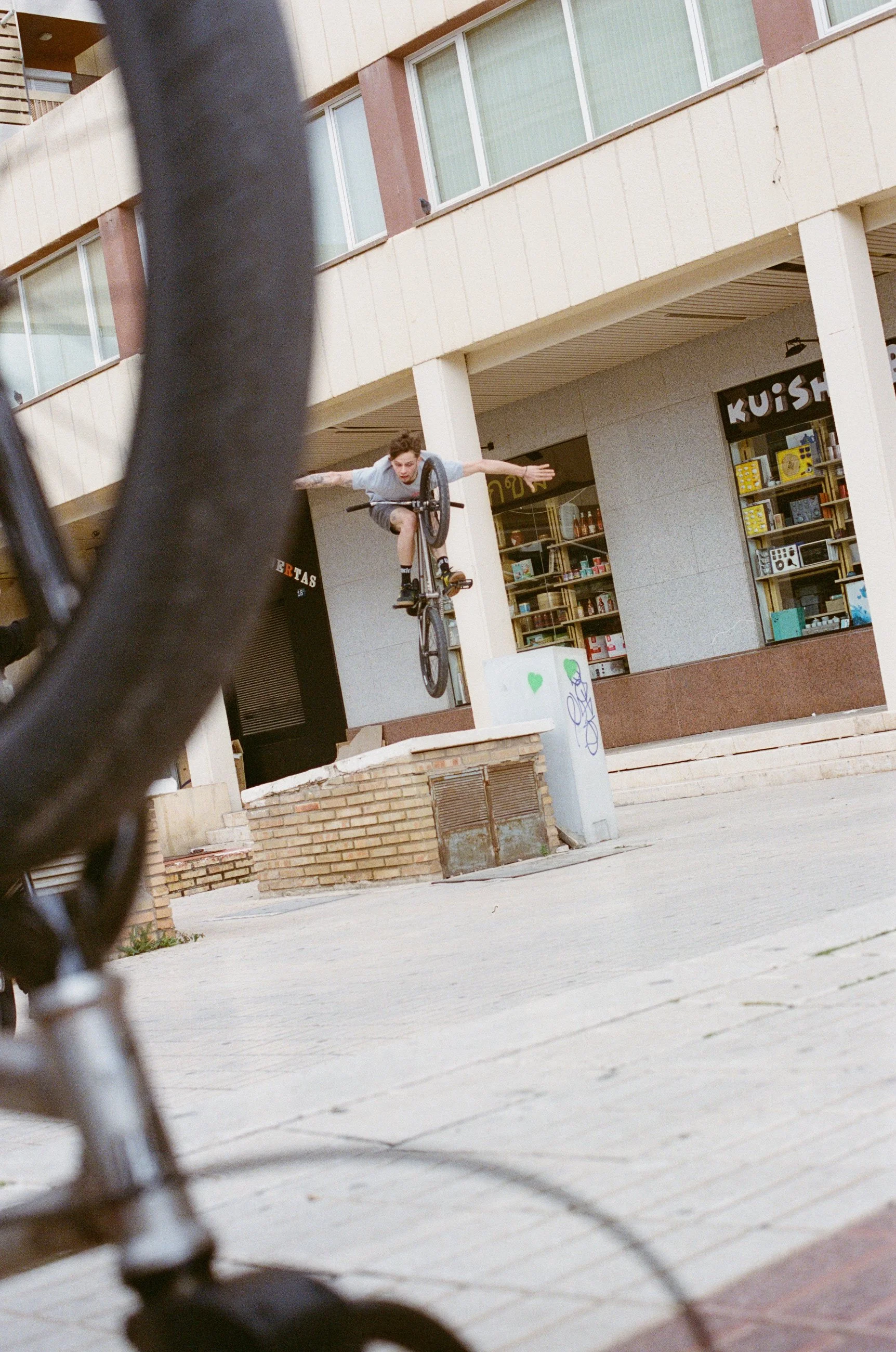 A person performing a bicycle stunt in an urban area, with part of another bicycle in the foreground, against a backdrop of storefronts and a multi-story building.