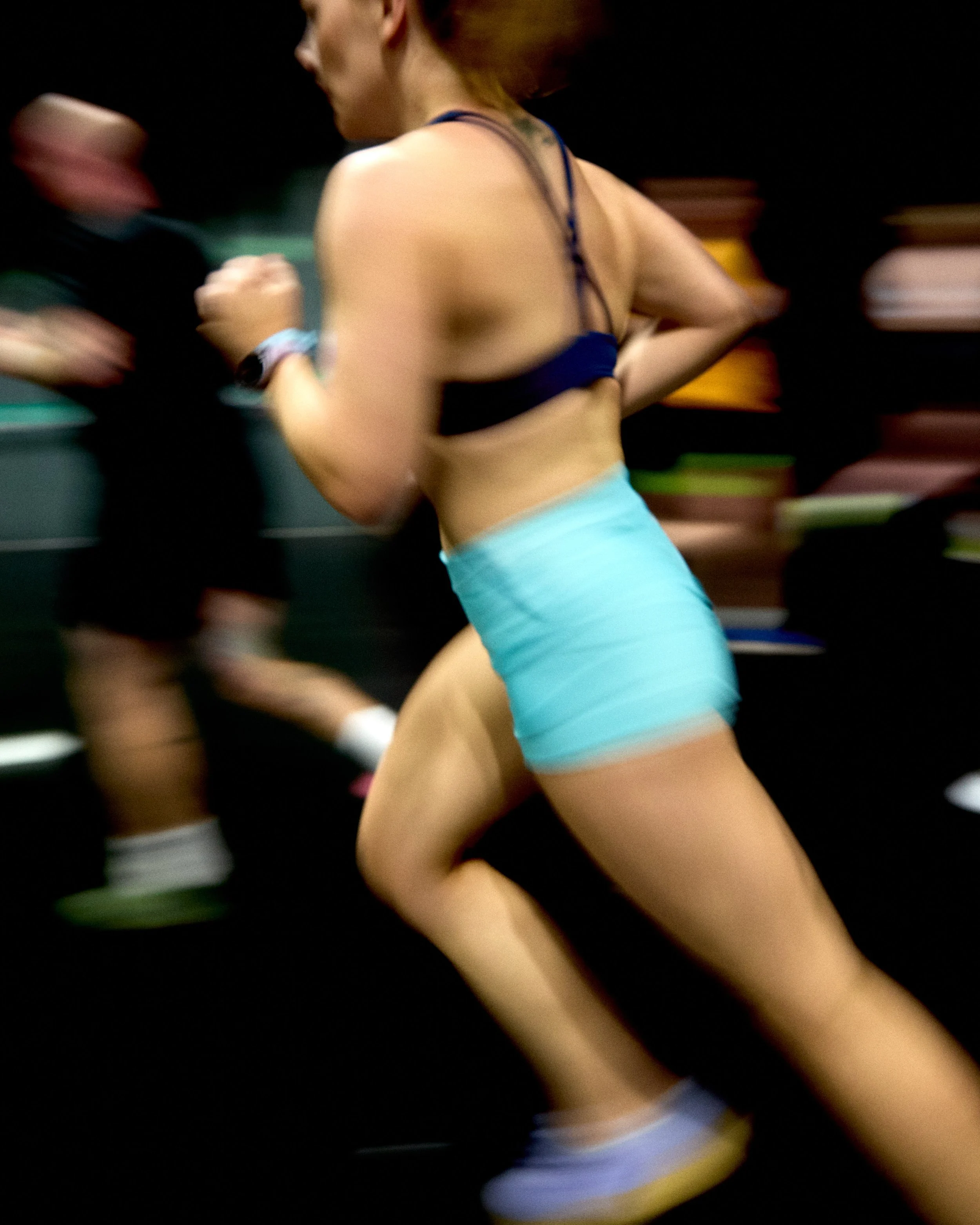 A woman running in a gym, wearing a sports bra and light blue shorts, with motion blur indicating fast movement.