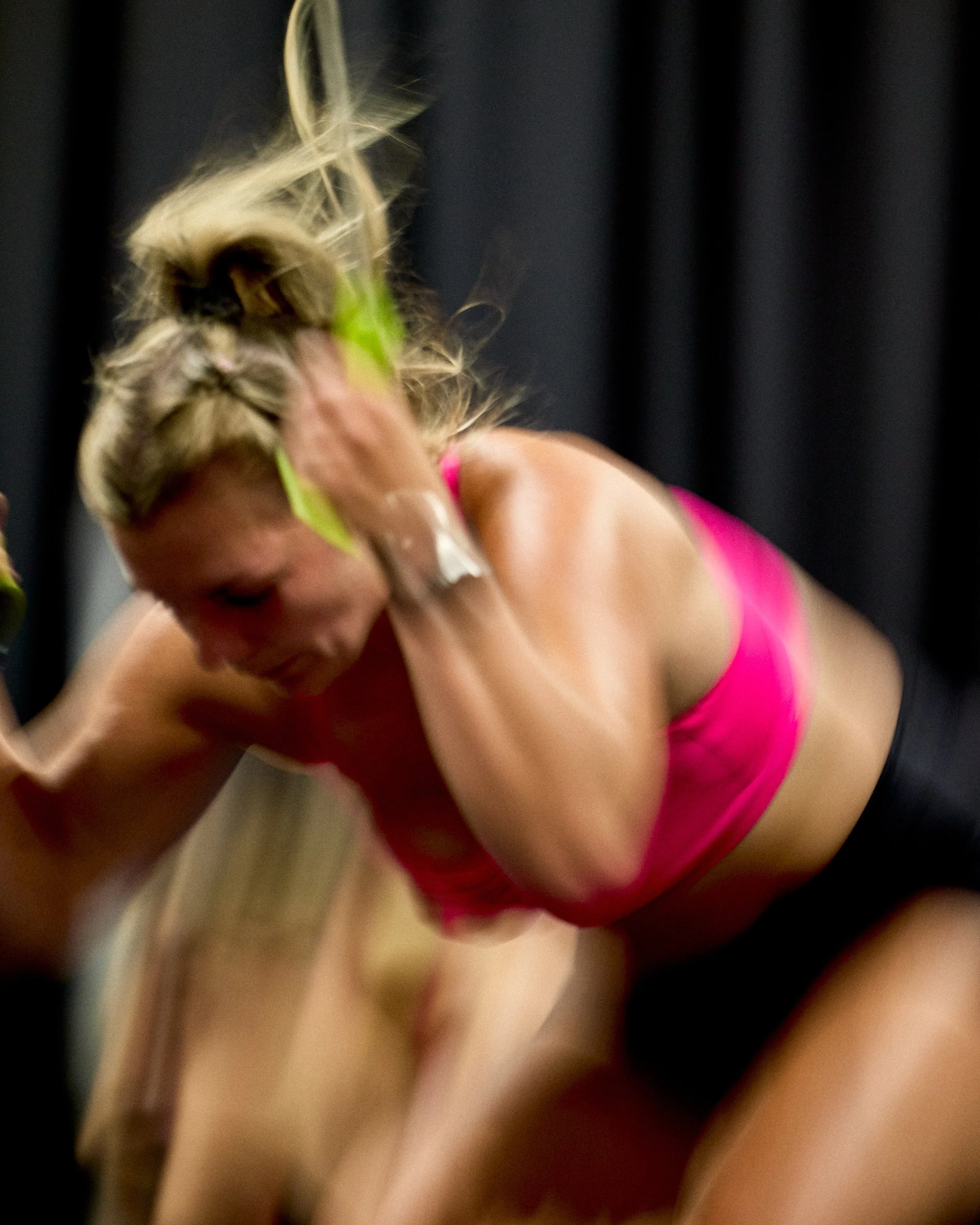 A woman with blonde hair, wearing a pink sports bra, engaged in a wrestling match on a black mat with a black curtain in the background.