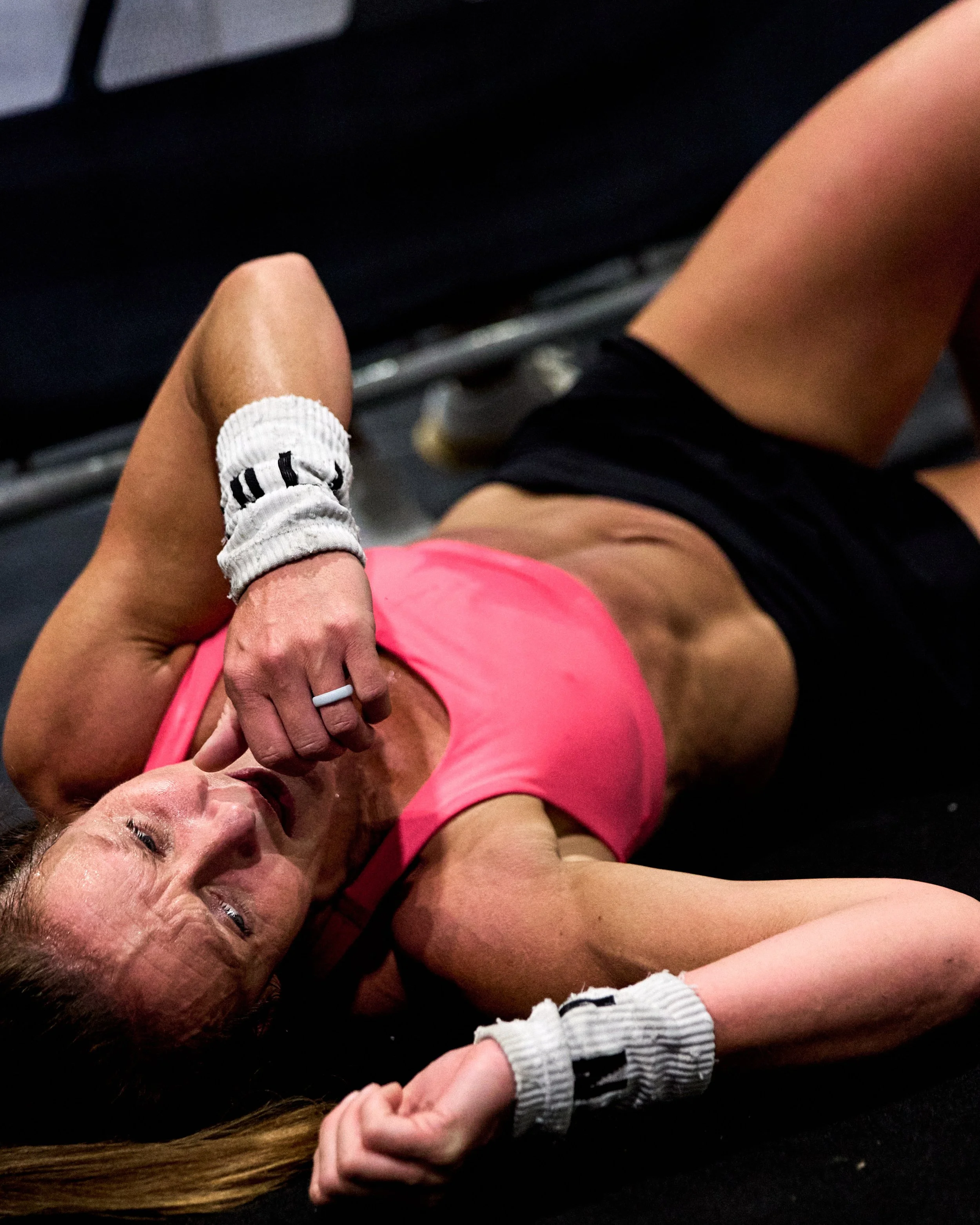 A woman lying on the floor after a workout, wearing a pink sports bra, black shorts, and white wristbands, with her left hand near her face and her right arm extended.