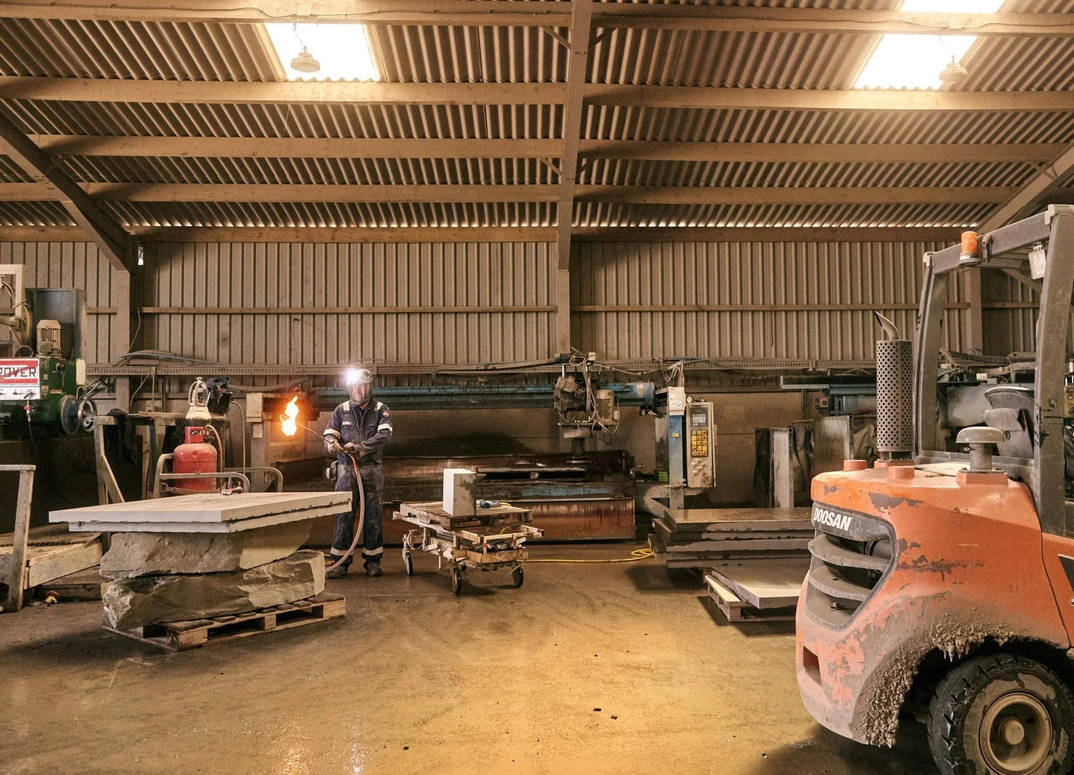 A worker using a blowtorch in an industrial warehouse with large machinery and equipment, including a forklift.