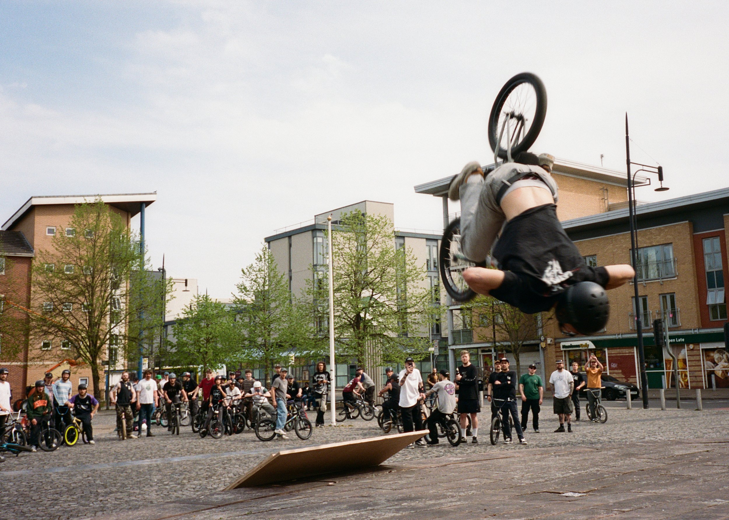 A person performing a flip on a BMX bike in an urban plaza, with a crowd of onlookers and modern buildings in the background.