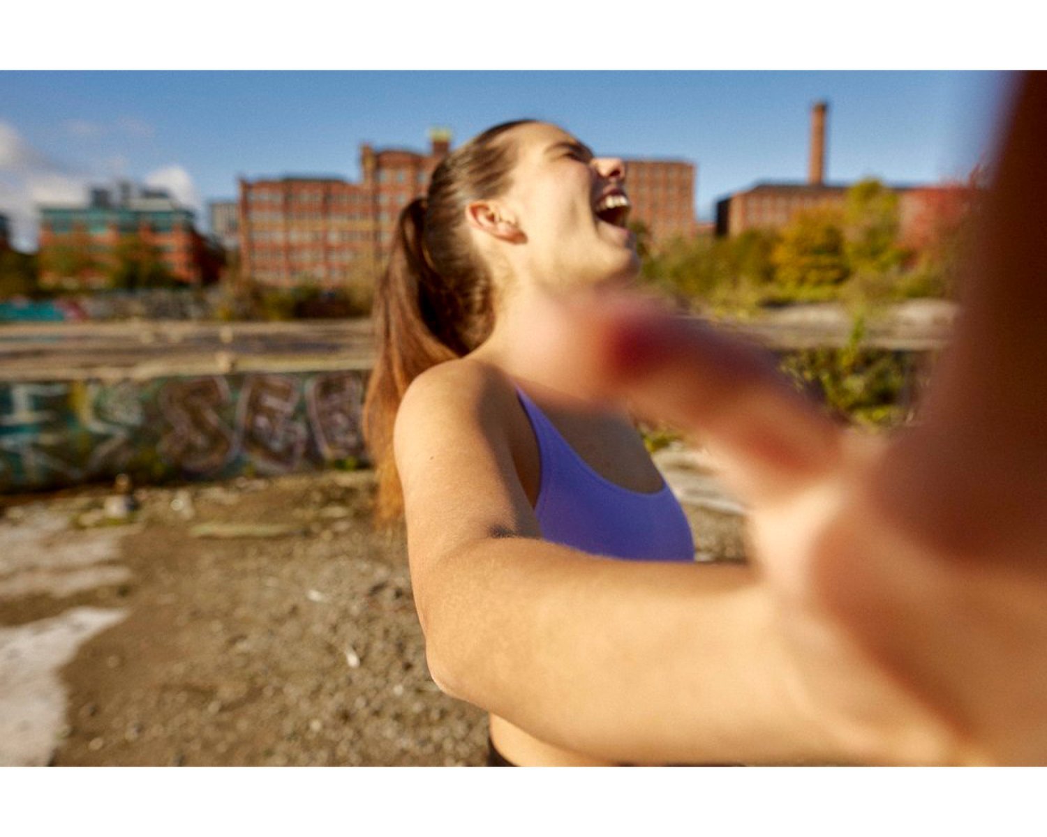 A woman with long hair wearing a blue sports bra, smiling and laughing outdoors on a sunny day with a cityscape and colorful graffiti in the background, as someone reaches toward the camera.