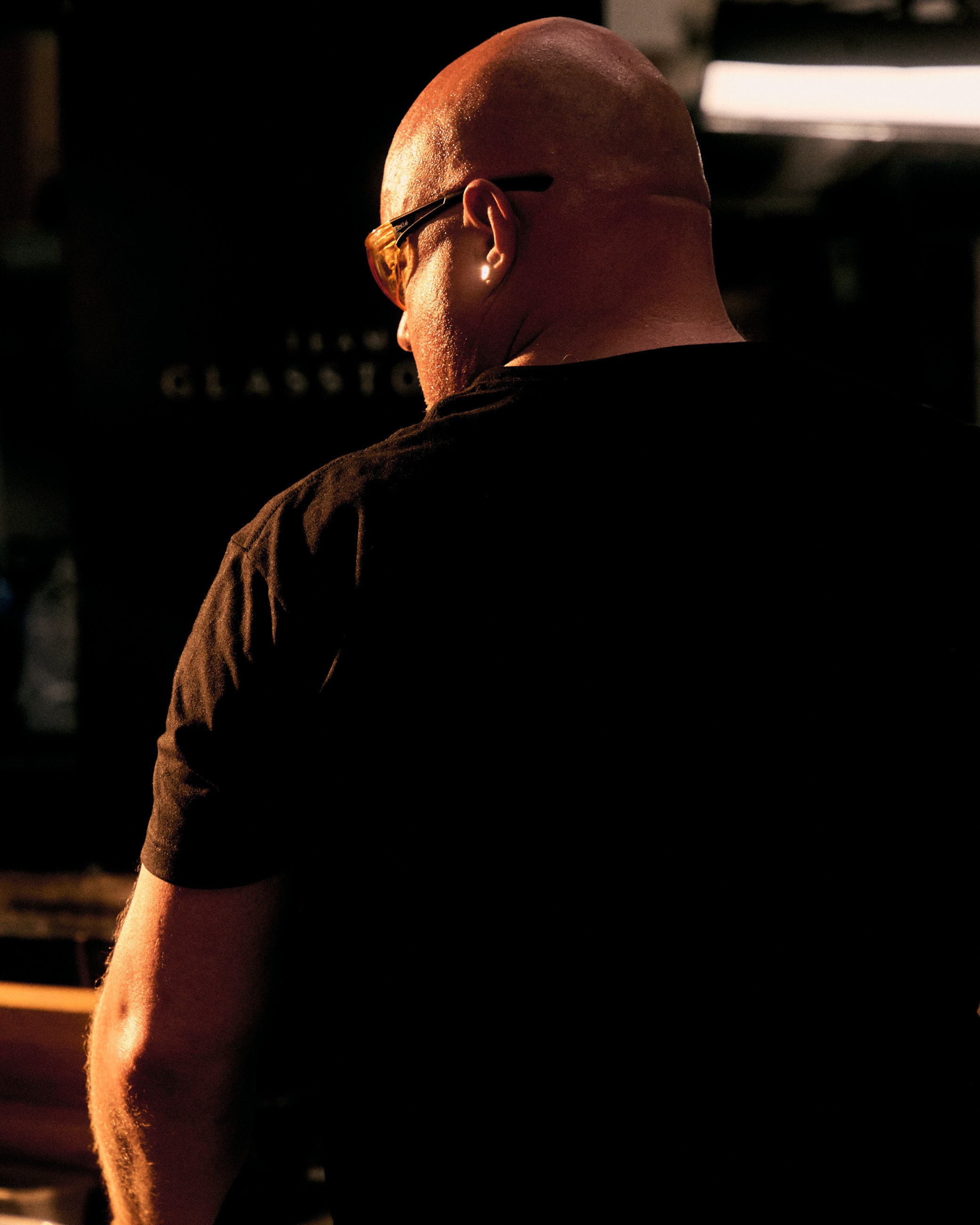 A man with a shaved head and glasses, facing away from the camera, in a dark environment with a warm light shining on his head and shoulder.