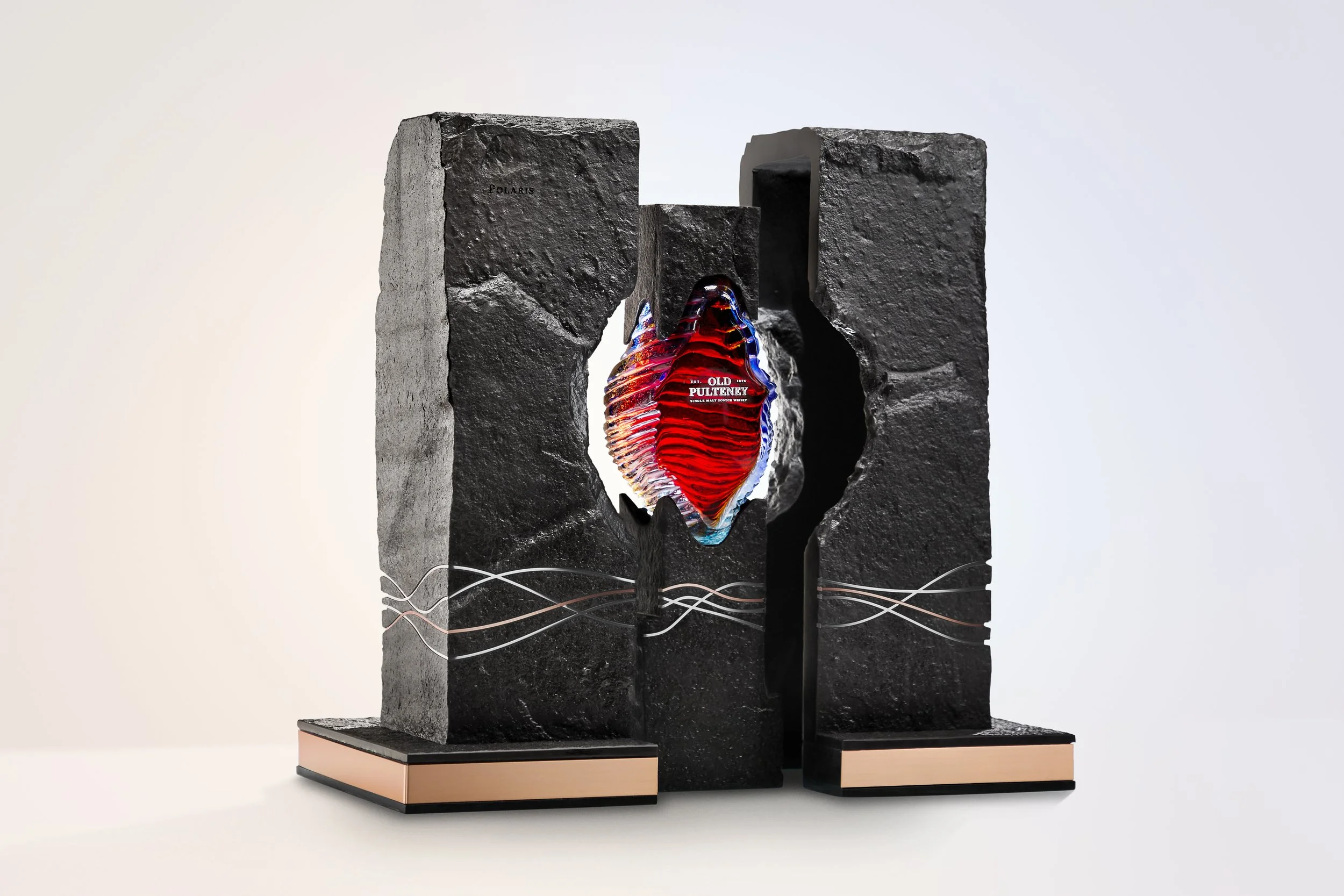 A modern sculpture features two large black textured stone blocks with a central hole, through which a glass sculpture of a red human heart is visible, with the label "Old Pulteney" and ripple-like patterns on the heart, mounted on a peach and black 