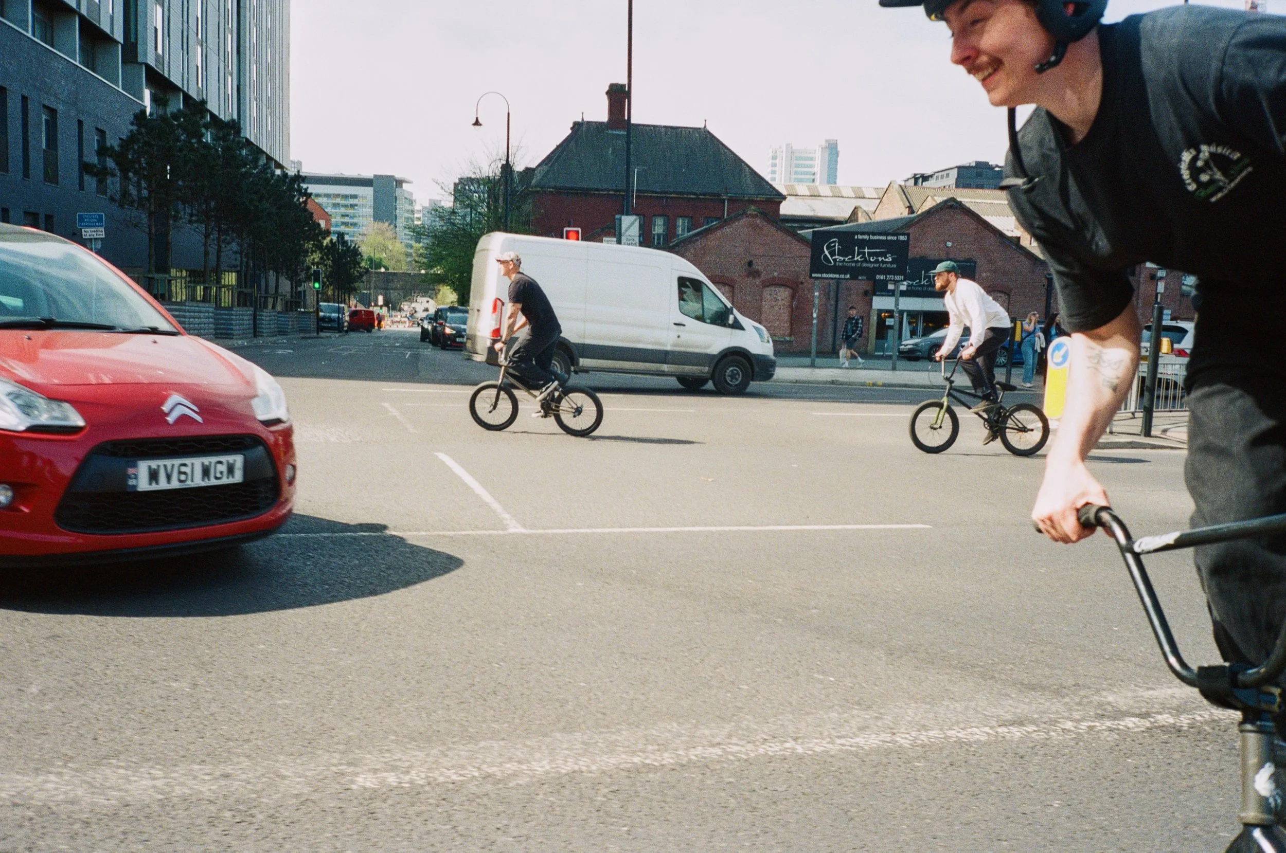 People riding BMX bikes on a city street at an intersection. The scene includes a red Citroën car, a white van, and a background of urban buildings and a brick house.