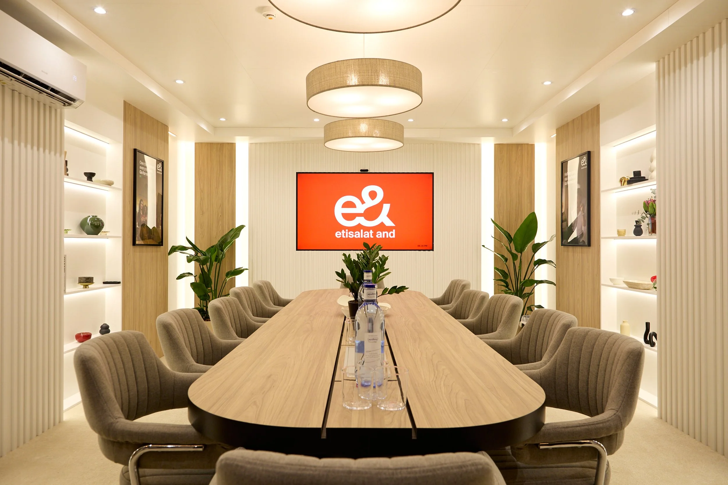 Modern conference room with a long wooden table, gray cushioned chairs, and a large red display screen showing Etisalat and e& logos. Decor includes potted plants, framed pictures, and decorative shelving with vases and art objects.