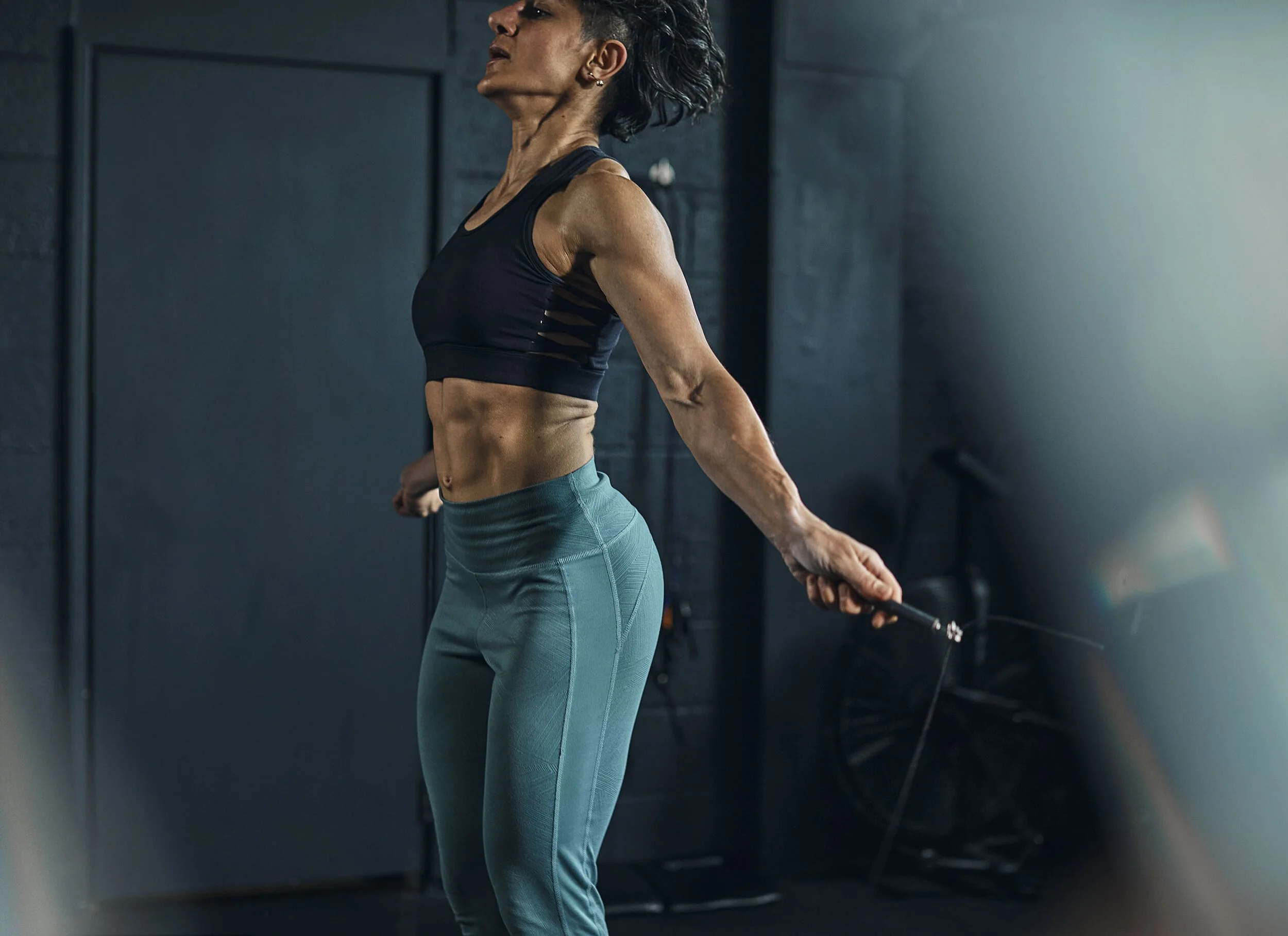 A fit woman with dark hair in a workout outfit is jumping rope in a gym, with dark walls and exercise equipment in the background.