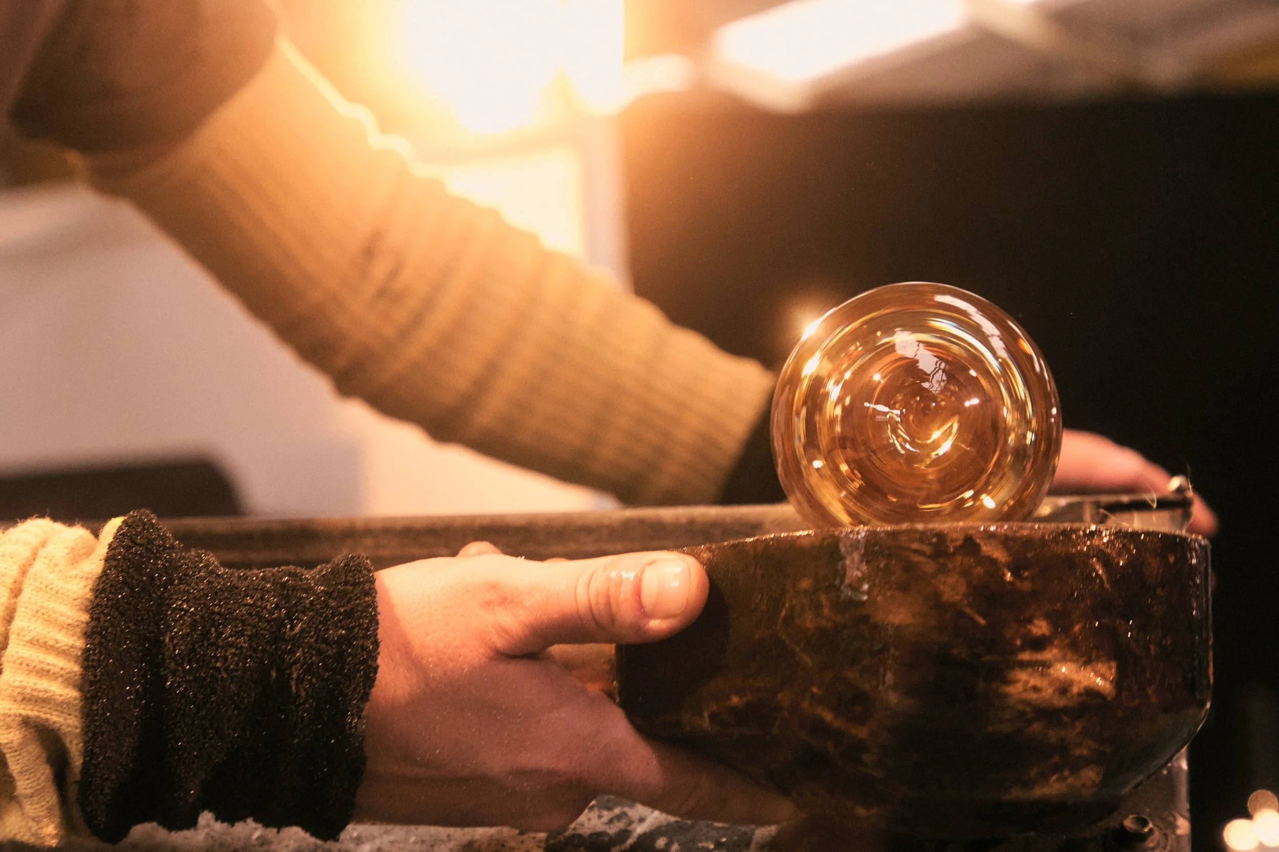 Person holding a glass of whiskey over a dark bowl, with sunlight in the background.