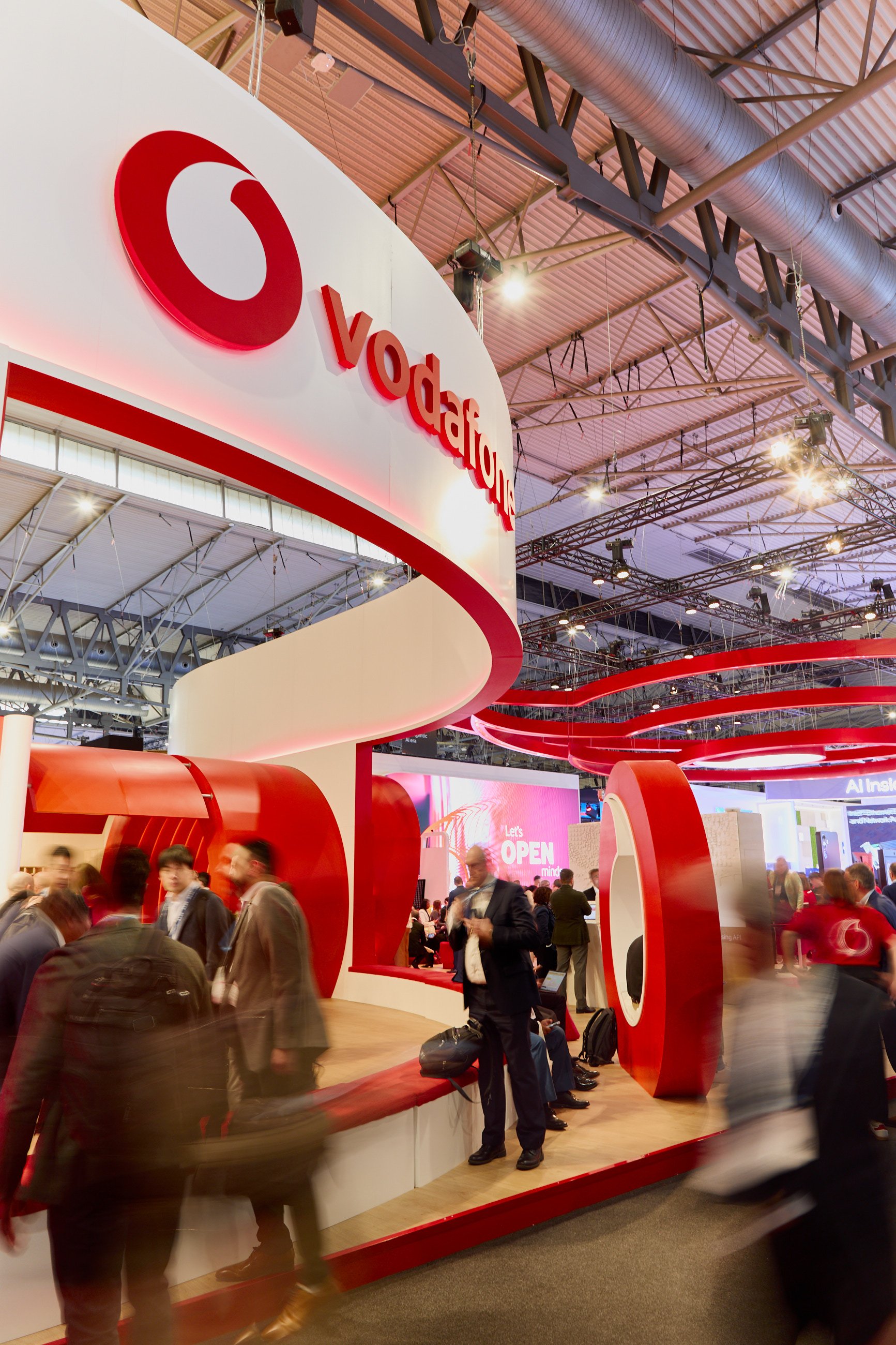 Vodafone booth at a trade show or convention center with a large red and white logo and curved architectural design, with people walking around.
