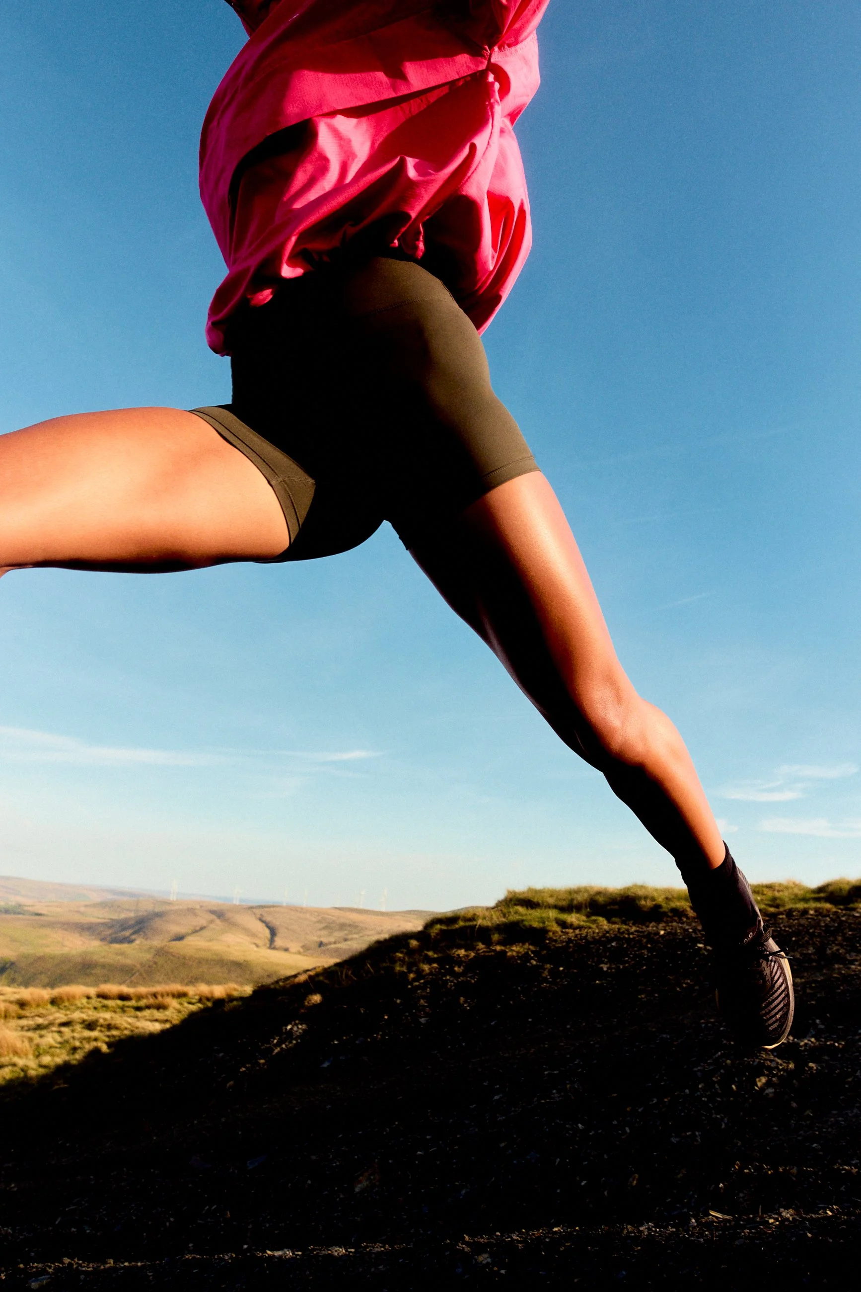 A person wearing a pink jacket and black athletic shorts running outdoors on a trail with hills and wind turbines in the background under a clear blue sky.