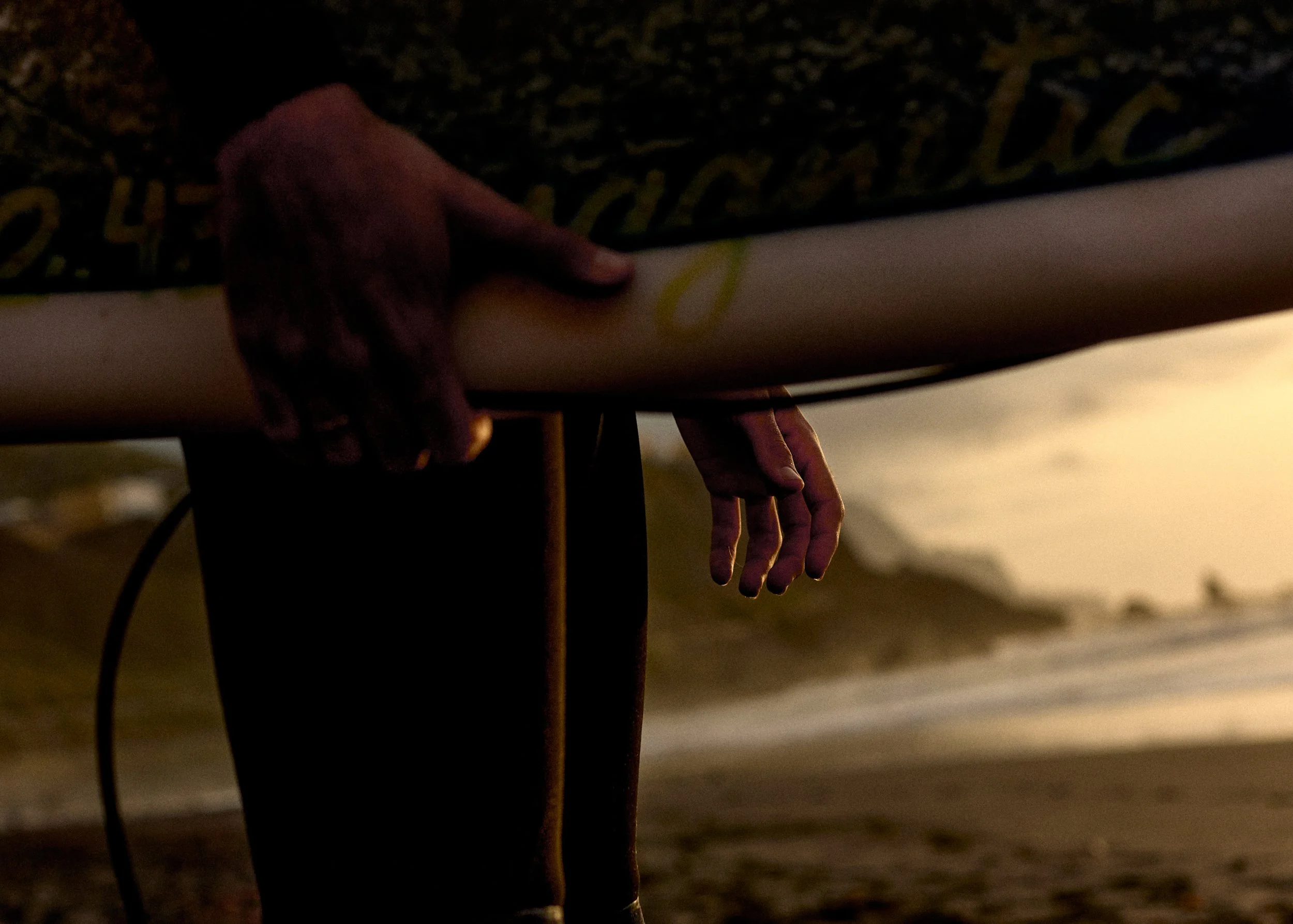 Close-up of a person’s hand gripping a surfboard at the beach during sunset, with the ocean and shoreline in the background.