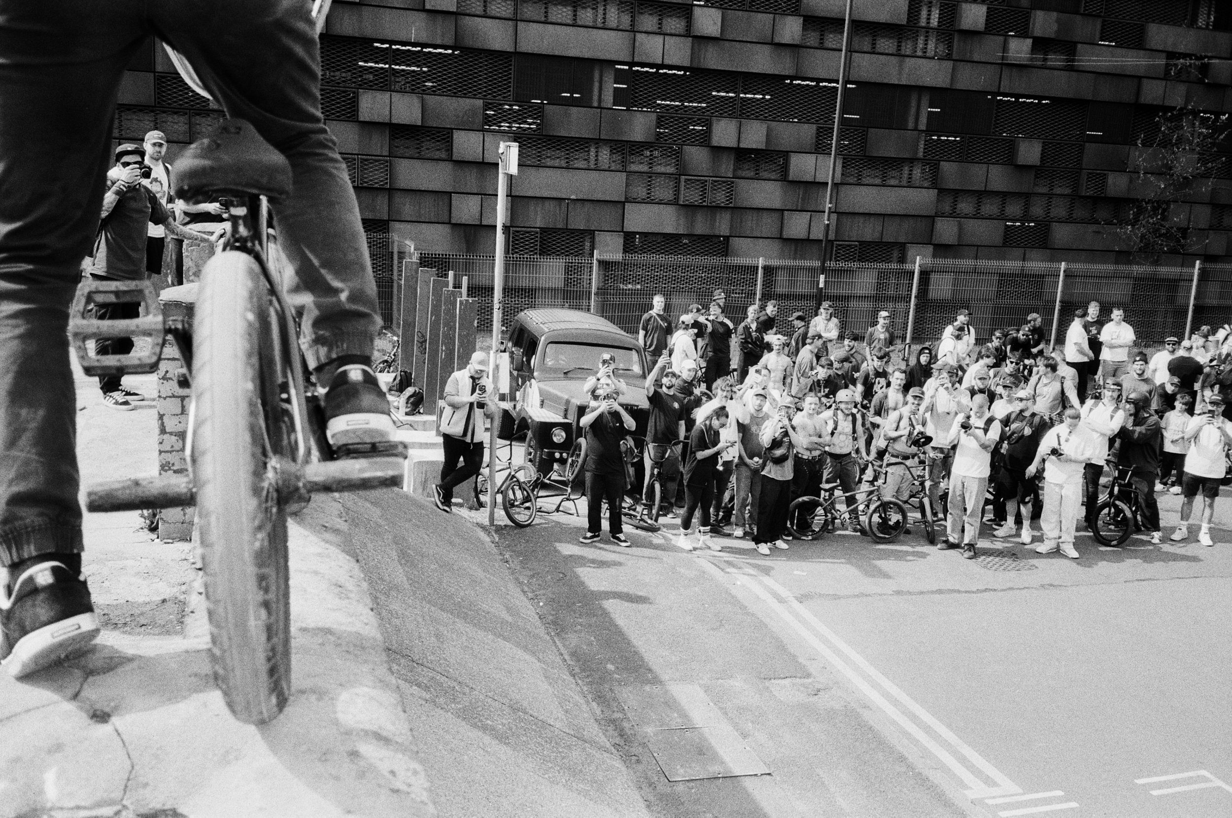 A black and white photo of a crowded urban street with many people holding phones and bicycles, as a cyclist rides on a ramp in the foreground.